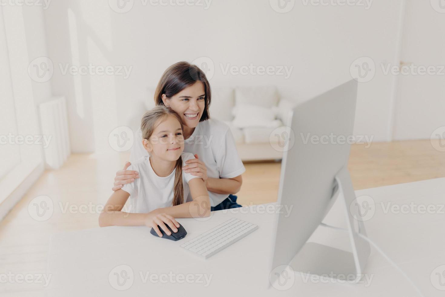 Indoor shot of happy lovely mother and daughter watch funny cartoon on computer, have pleasant smiles, enjoy togetherness, spend free time or weekend at home, spacious light empty living room photo