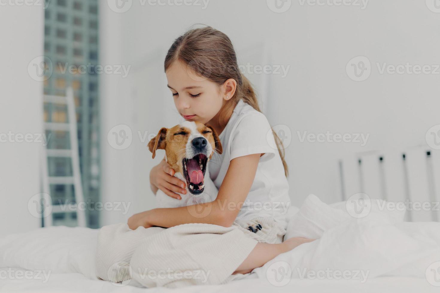 Fair haired small girl with pony tail wears casual pyjamas, embraces dog who yawns, pose together on comfortable bed, expresses love and care to domestic animal, spends morning weekend at home photo