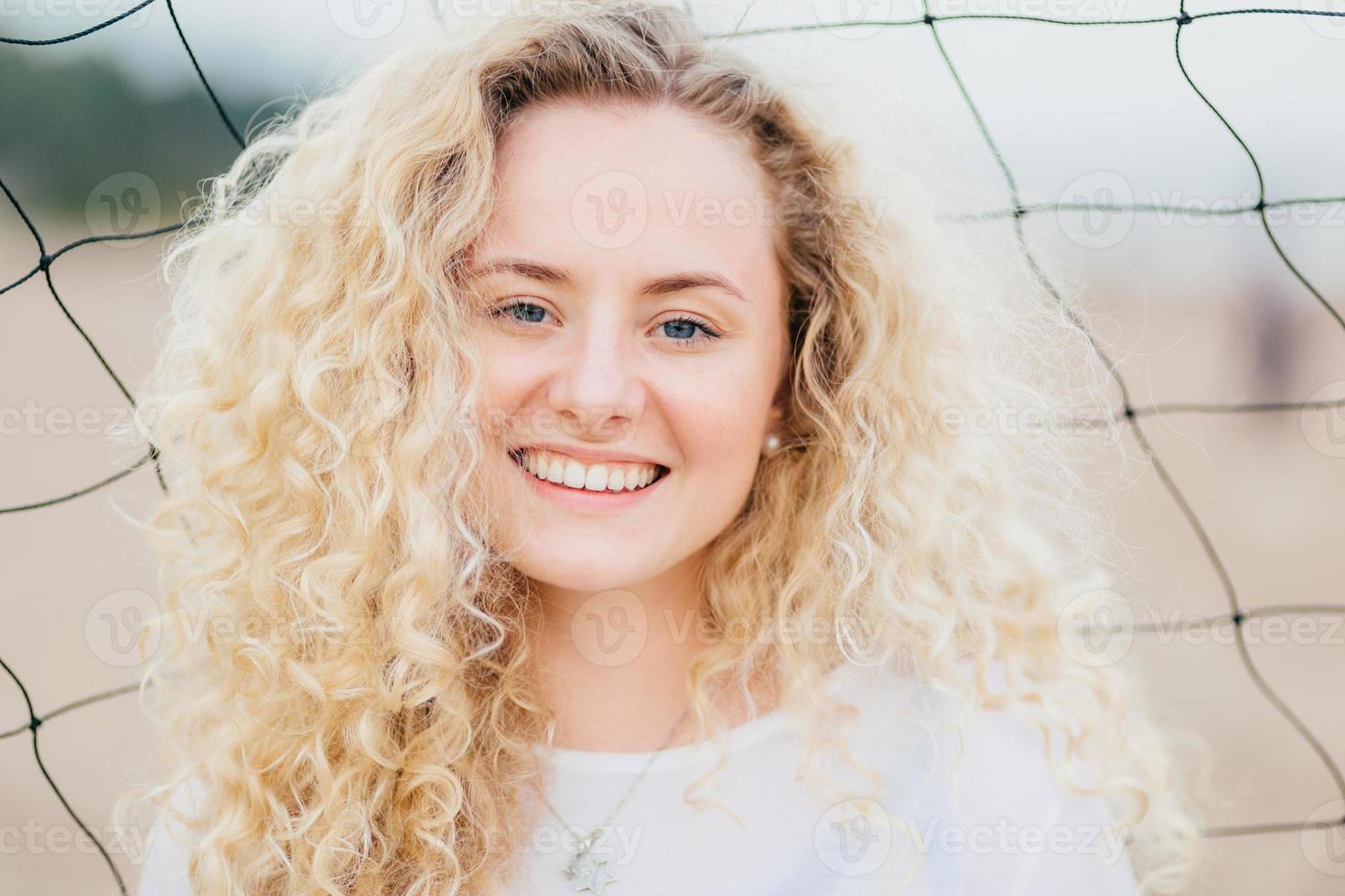 Lovely cute young woman has fresh skin, curly light hair, smiles gladfully, dressed in casual clothes, stands near tennis net, being satisfied to recieve compliment. People, happiness concept photo