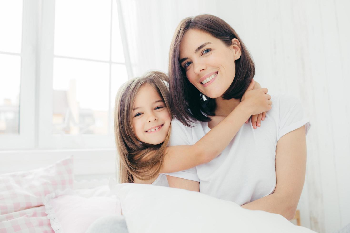 la hija positiva y la madre se abrazan, están animadas después de un buen sueño, posan en el dormitorio, sonríen suavemente en la cara, tienen una apariencia agradable. niña bonita con su madre. mañana y hora de acostarse foto