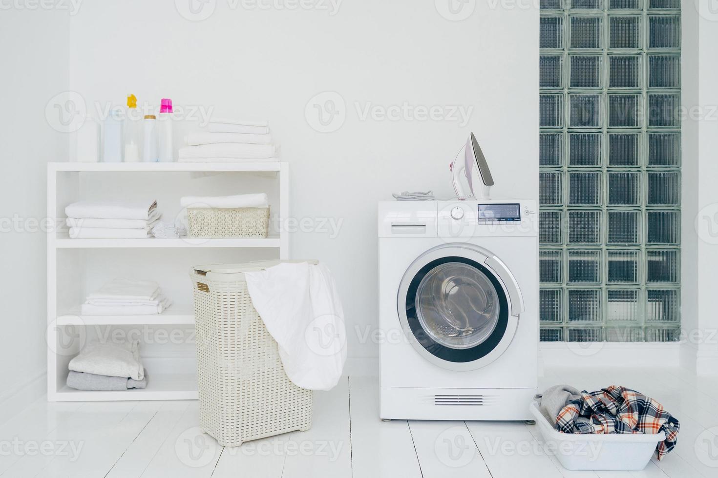 Laundry room with washing machine, dirty clothes in basket, iron and little shelf with neatly folded linen. Domestic room interior. Washing concept photo