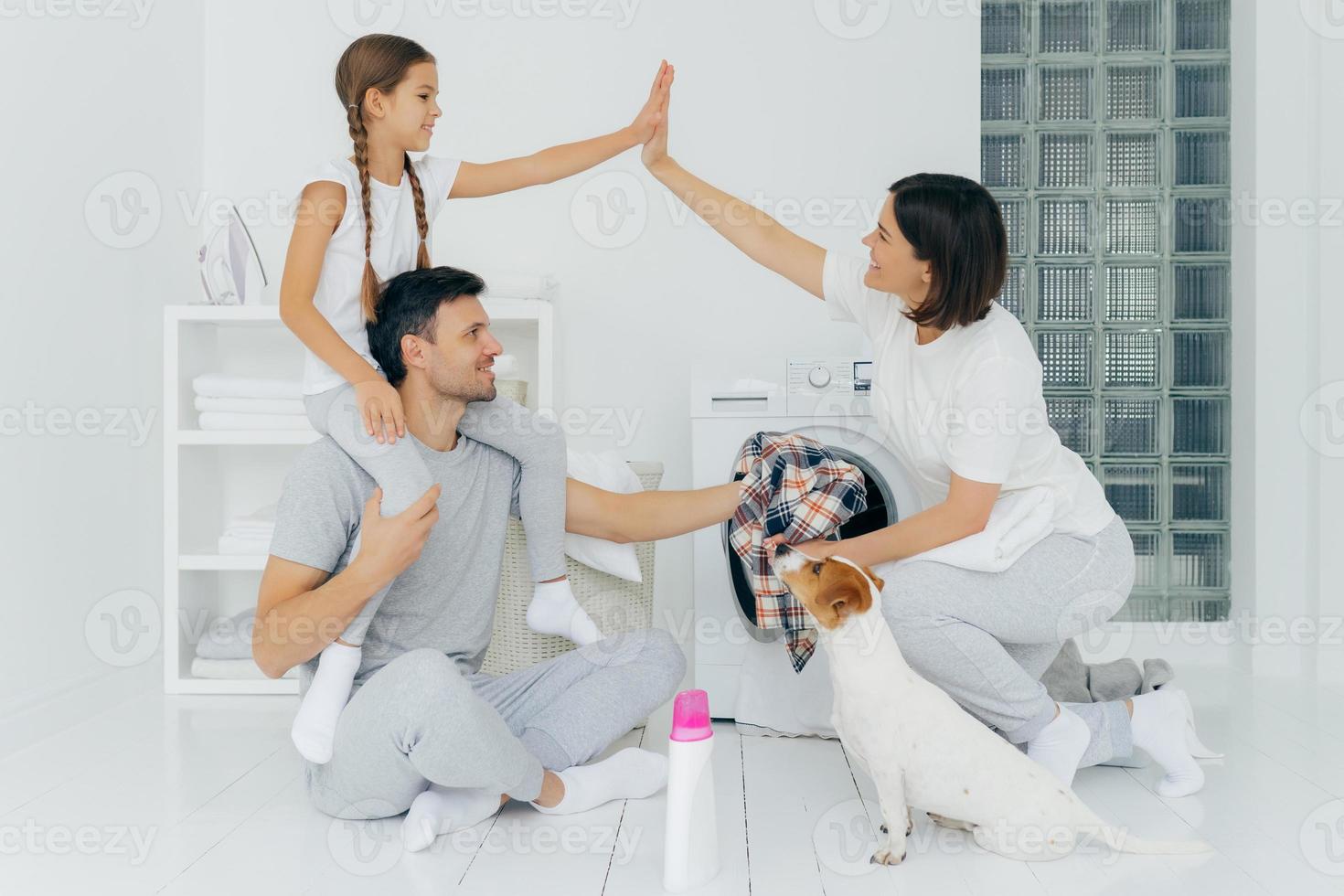 Well done. Daughter and mother make high five, rejoice almost finishing domestic work, girl rides on dads shoulders, wash clothing in laundry room with white walls. Jack russell terrier looks at host photo