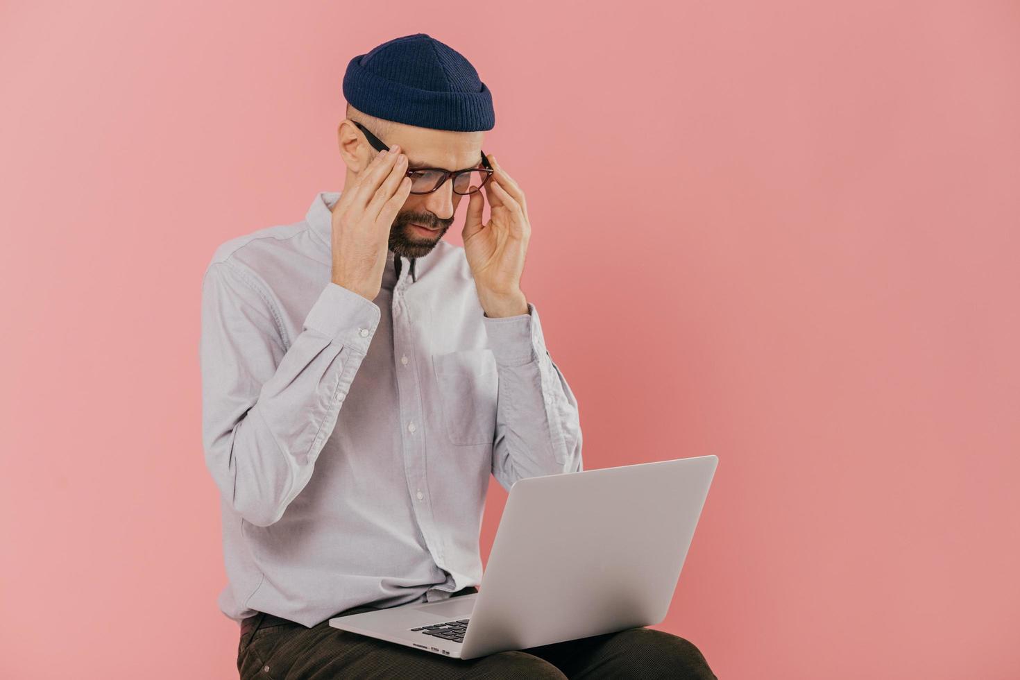 Thoughtful serious student keeps hands on spectalces, has online conversation via portable laptop computer, watches webinar, wears hat and white shirt, models over pink background with free space photo