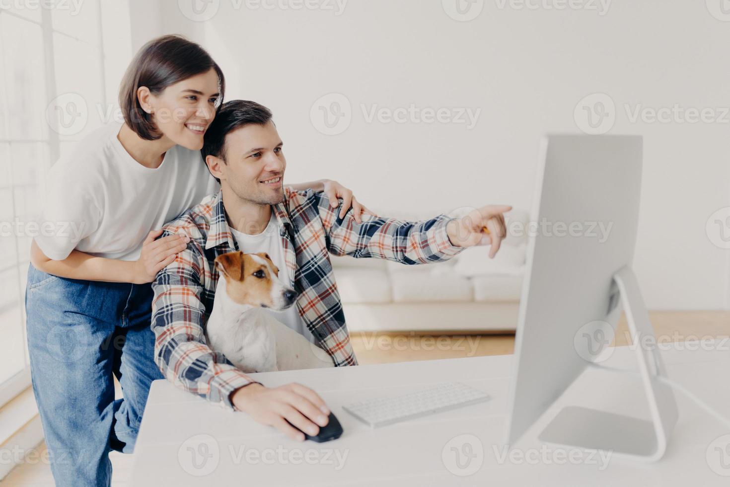 Affectionate brunette wife supports husband with doing project work, happy man points into monitor, tells something, shares his ideas, works together with pedigree dog, pose in spacious cabinet photo