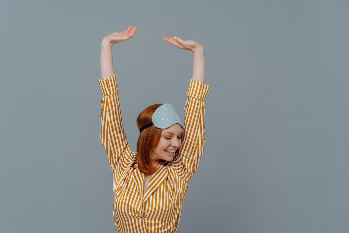 Feeling rechared. Beautiful freckled girl keeps arms raised up, feels total relaxation, smiles positively, wears sleepmask and pajamas, isolated over grey background. Rest, sleep and bedtime concept photo