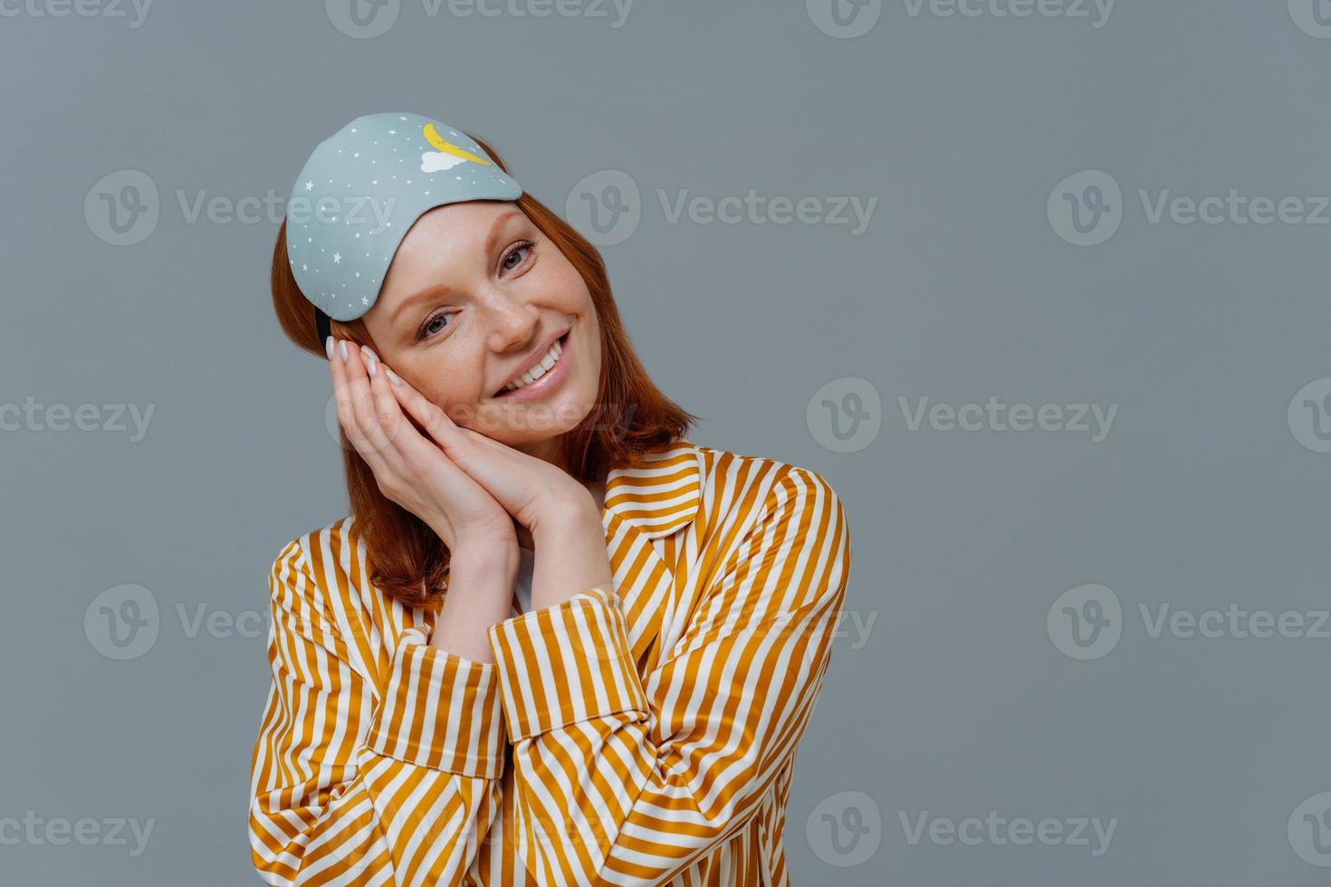 Horizontal shot of attractive ginger woman feels glad after having enough sleep, just woke up, tilts head on hands, wears blindfold and pajamas, isolated on grey wall, empty space. Sleep gesture photo