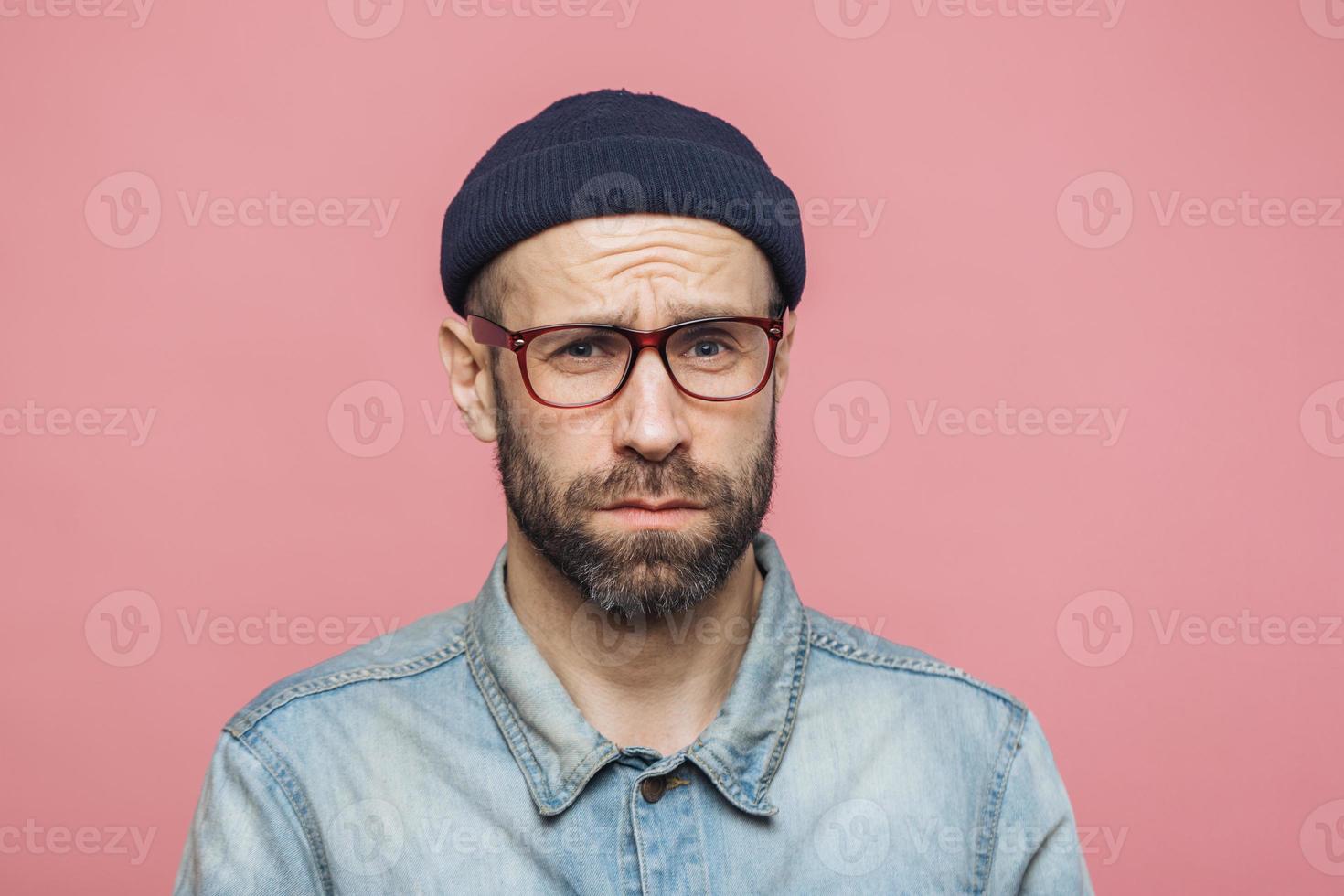 Grumpy bearded man with offended expression, being dissatisfied with something, frowns face, wears spectacles, hat and shirt, isolated over pink background. Displeasure and negativity concept photo