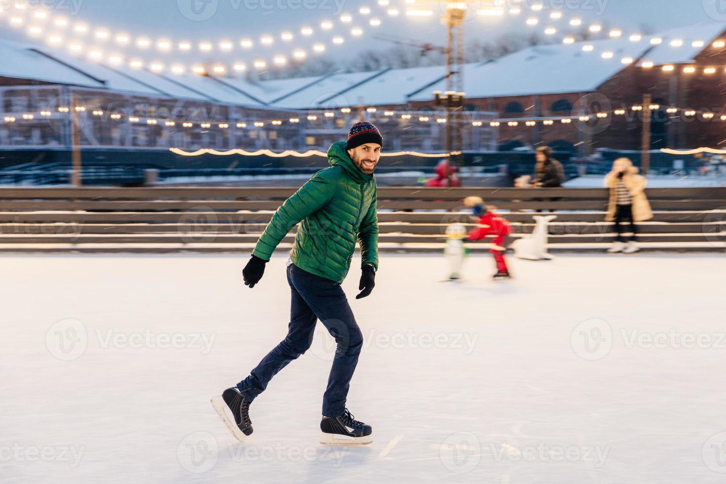 Professional male skater shows his skating talents, being sure on ice ring, looks delightfully in camera, demonstrates his nice skills. Smiling happy man on skating ring. Leisure and professionalism photo