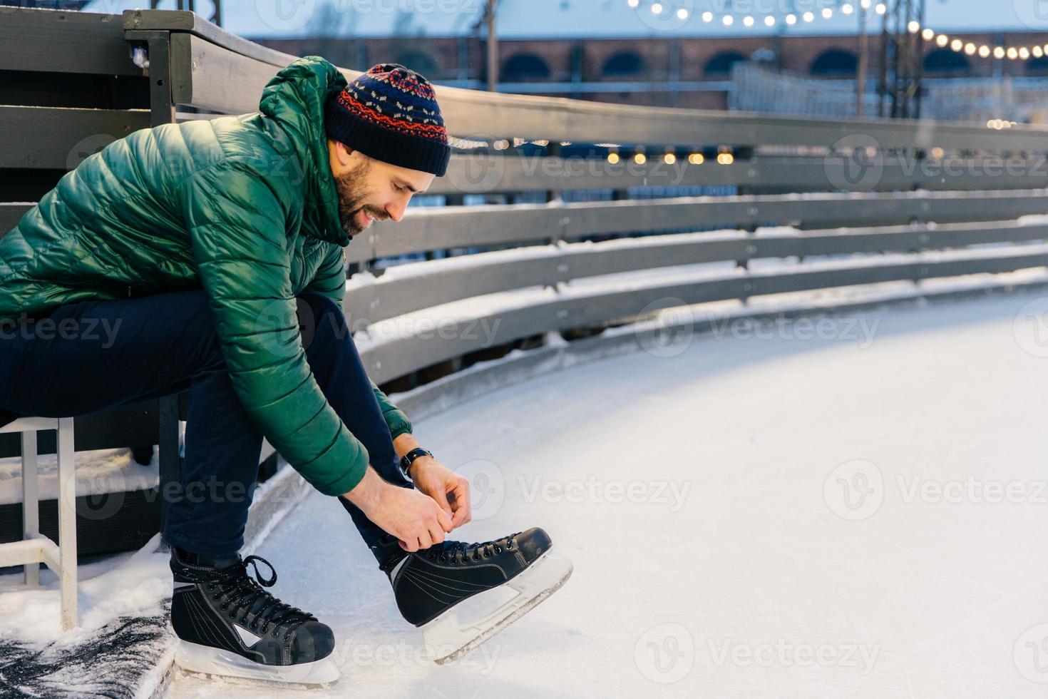 People, sport, winter, leisure concept. Smiling delightful male laces up skates, going to practice on ice ring, being in good mood, likes winter and go skating with best friends. Hobby concept photo