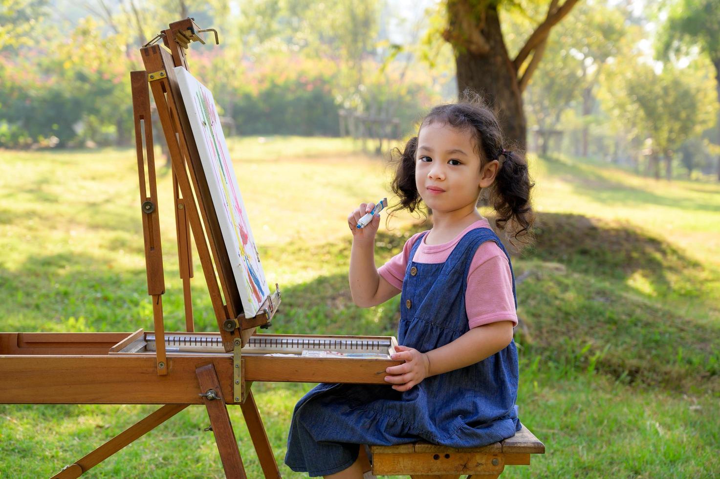 una niña pequeña está sentada en el banco de madera y pintada en el lienzo colocado en un puesto de dibujo foto