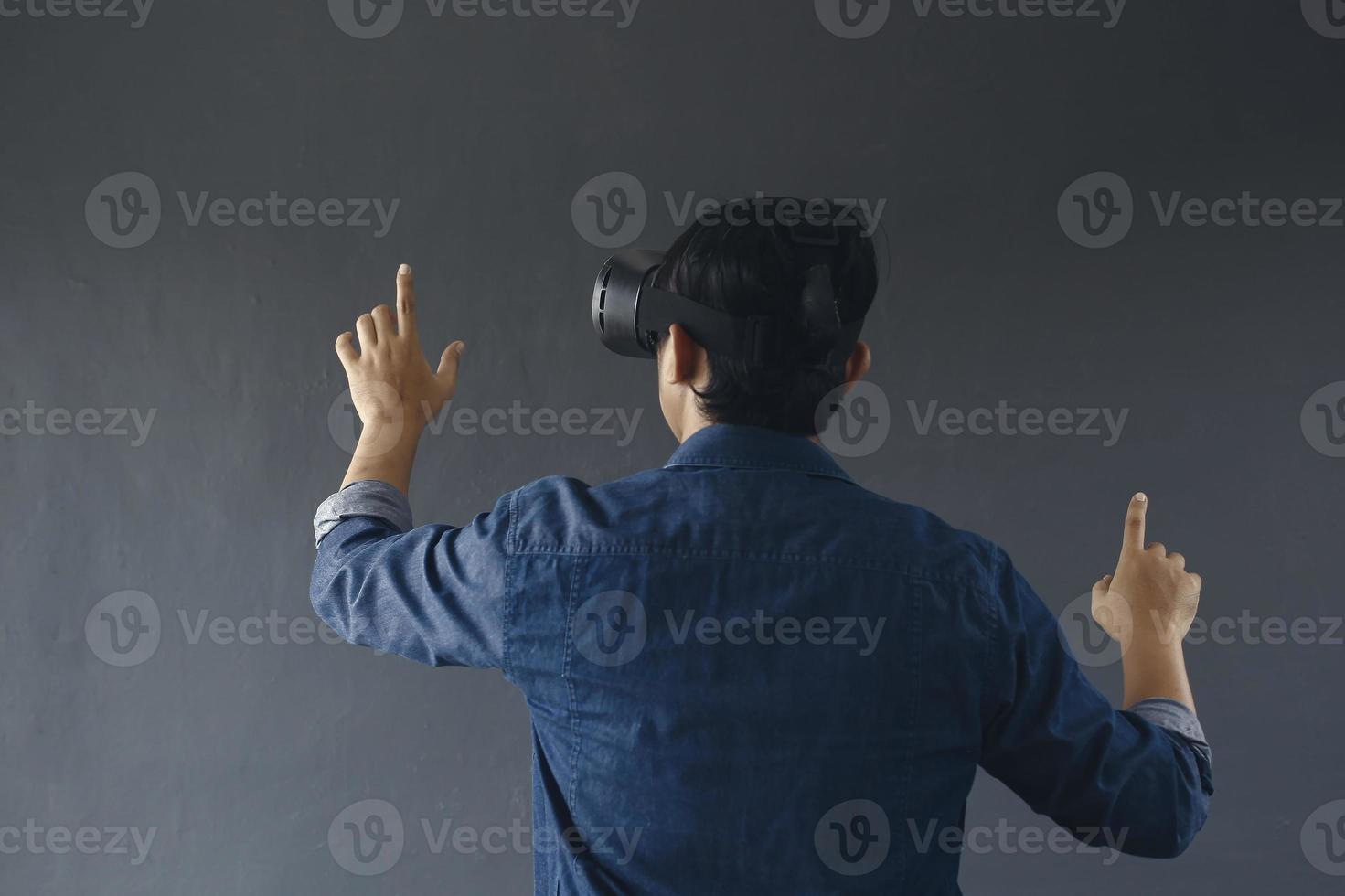 Back view of young man in casual style wearing virtual reality googles, his finger pointing out or touching blank screen. Copy space for futuristic games and education concept. photo