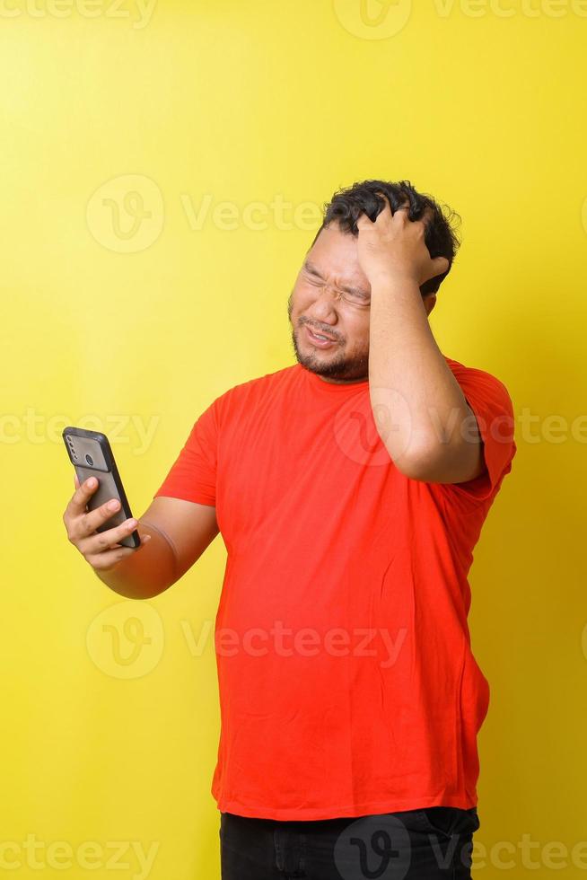 Asian fat man in red tshirt with sad expression and suddenly headache when holding smartphone isolated on yellow background photo