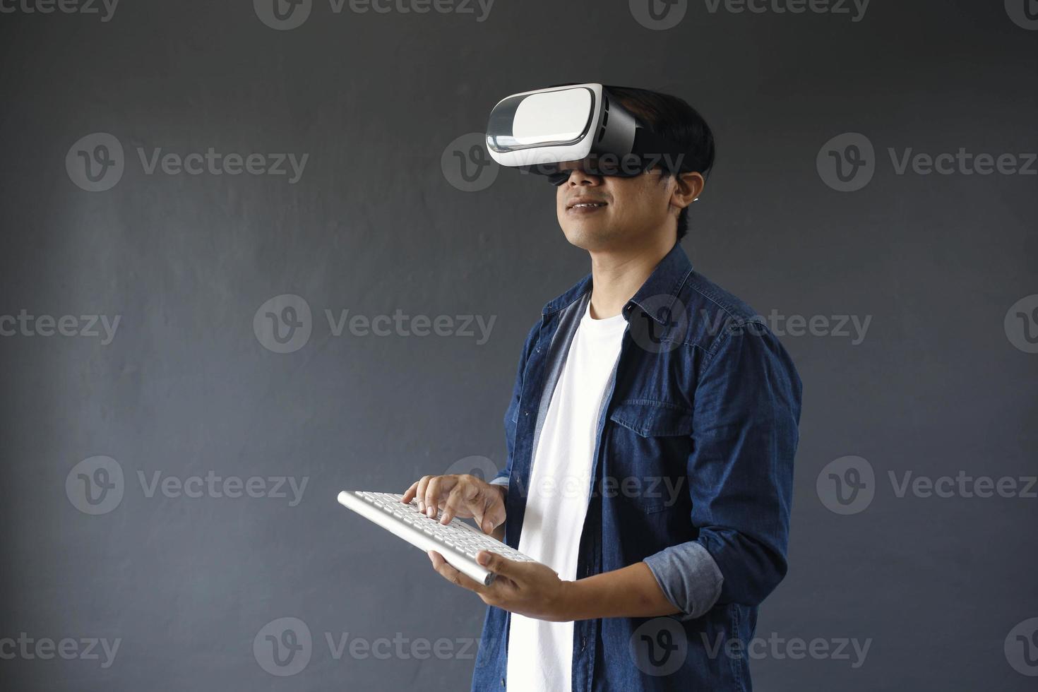 Young man in casual style using Virtual Reality glasses and wireless keyboard on dark grey background. Copy space for futuristic concept. photo