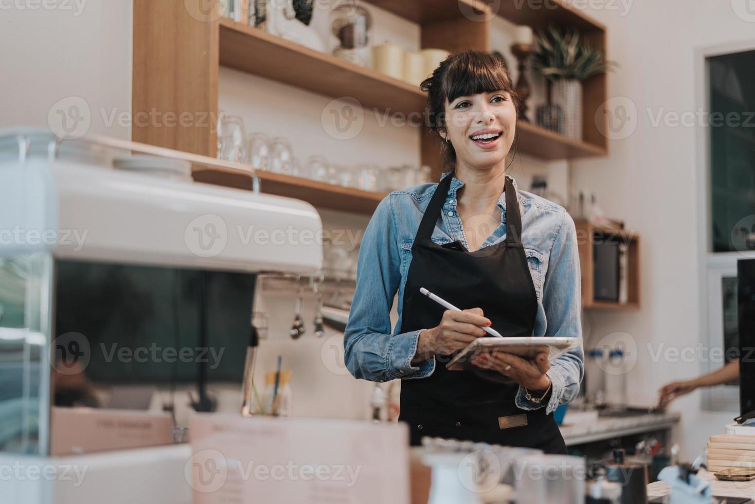 Happy woman coffee owner standing behind the counter of a coffee shop. female barista standing and holding digital tablet behind counter in cafe. Business owner concept. photo