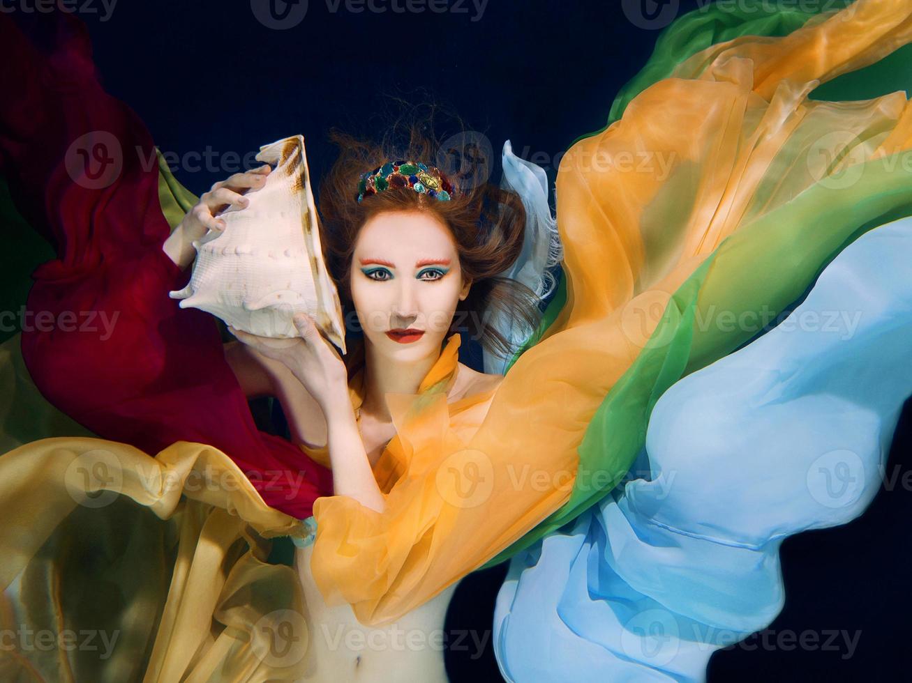 Redhead woman with big sea shell in colorful clothes on the dark background underwater photo