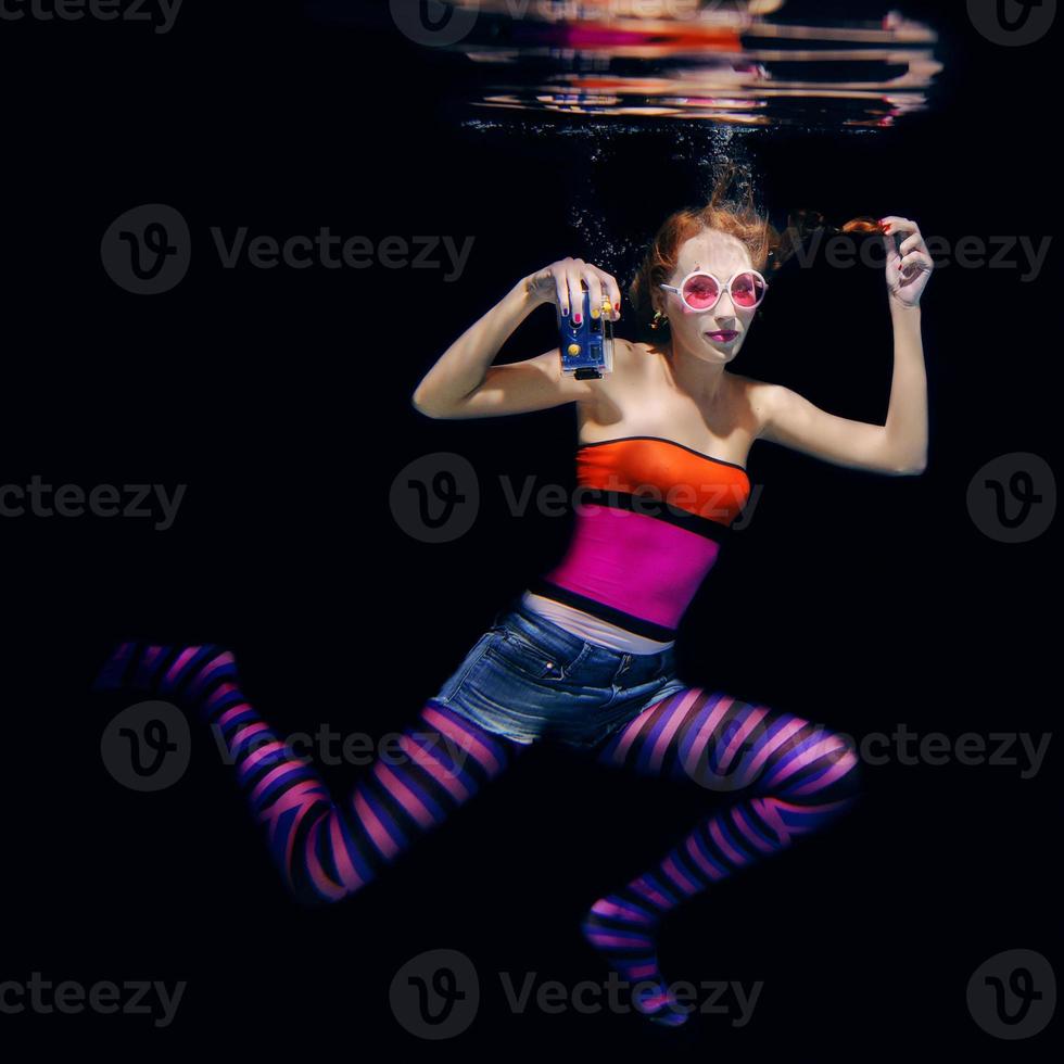 redhead funny woman in colorful clothes and pink sunglasses on the dark background swimming underwater with photo camera. Travel, sightseeing concept
