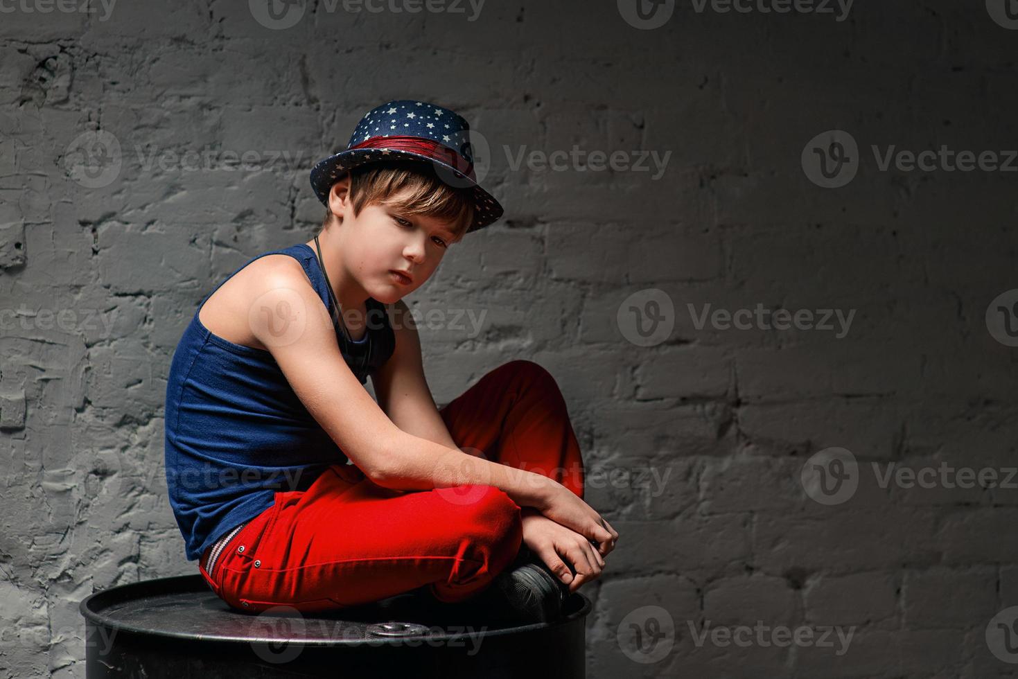 Portrait of cool young hip hop boy in blue hat and red pants sitting on black barrel in the loft photo