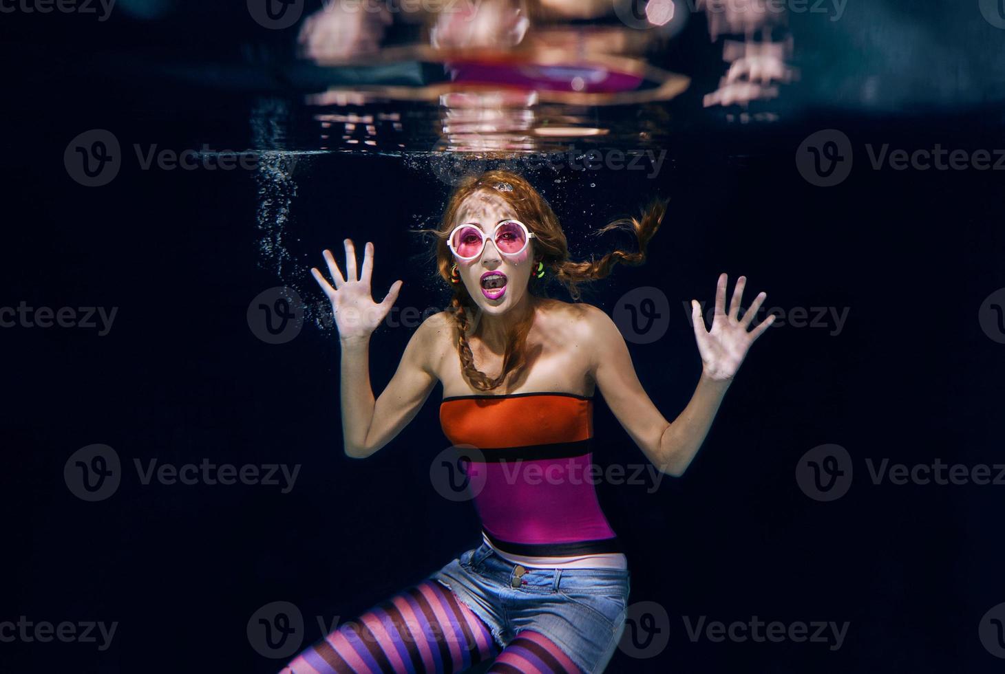 redhead funny woman in colorful clothes and pink sunglasses on the dark background swimming underwater photo