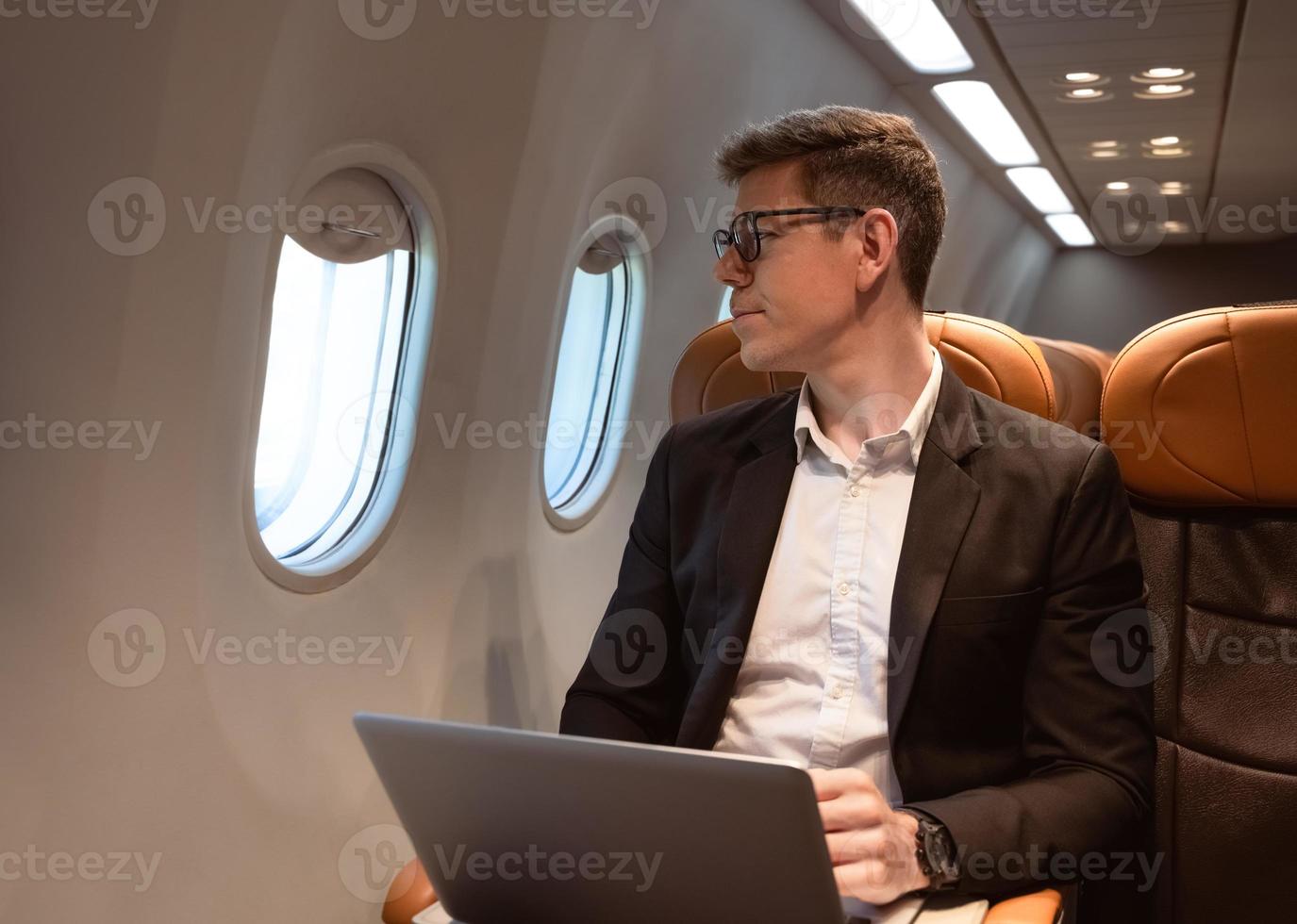 Businessman working with laptop while sitting in airplane seat and looking through aeroplane window. Business traveler concept photo