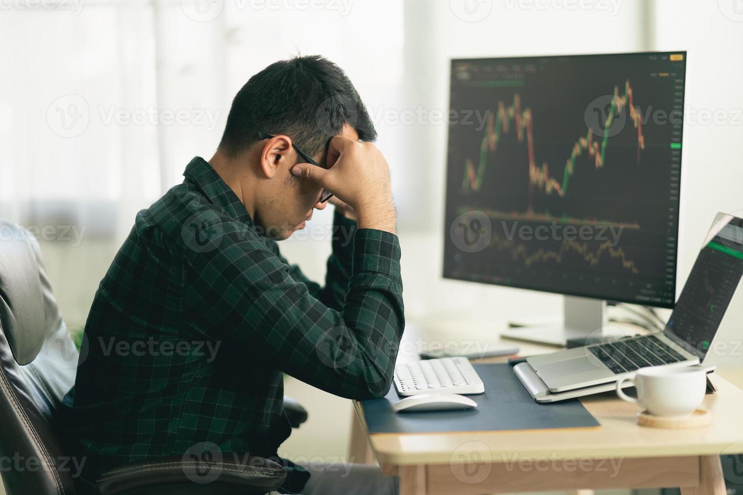 Asian man trader unhappy and serious sitting at home office in front of monitors with cryptocurrency graph holding smartphone monitoring cryptocurrency price photo
