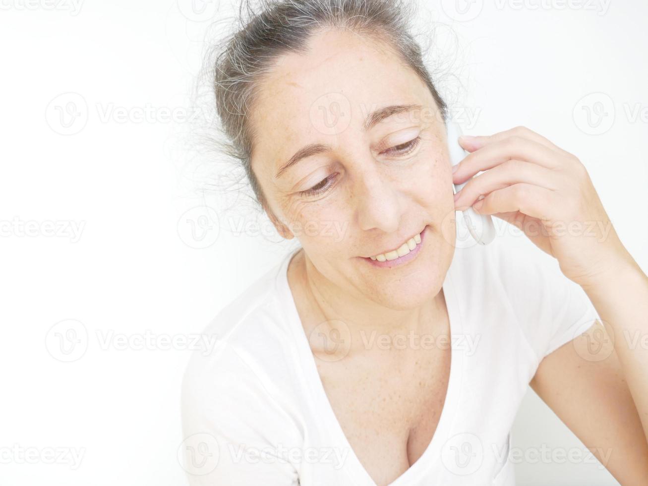mujer de cuarenta y nueve años con una camiseta blanca sobre un fondo blanco hablando por su celular foto