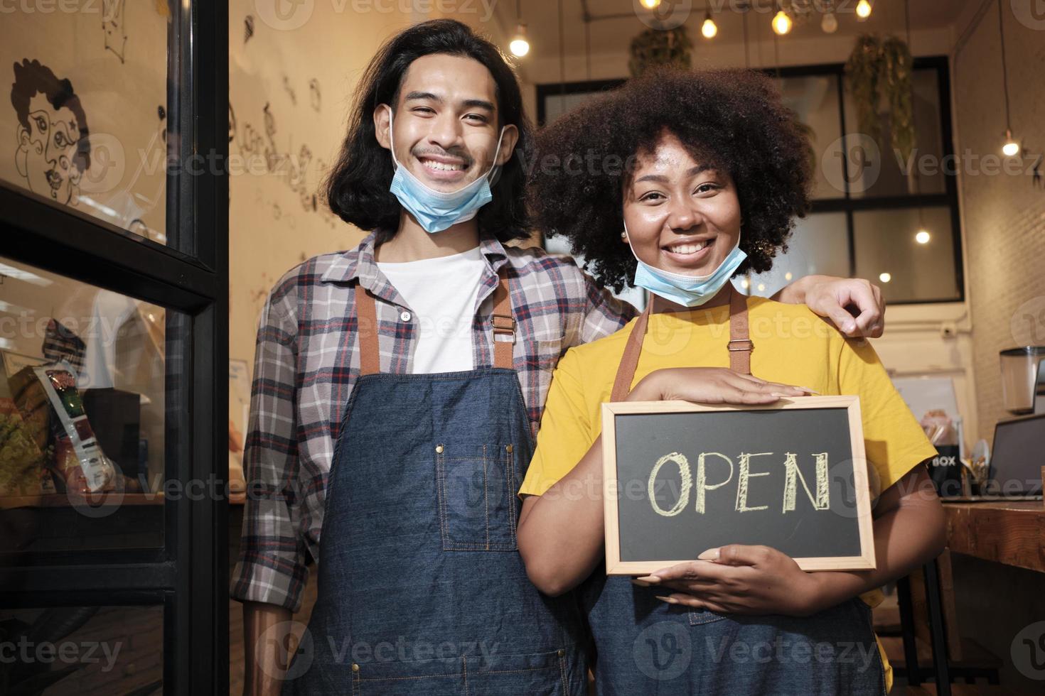 Two young startup barista partners stand at casual cafe door, write and show open sign before taking off face masks, happy and cheerful smiles with coffee shop service jobs after COVID-19 lifestyle. photo