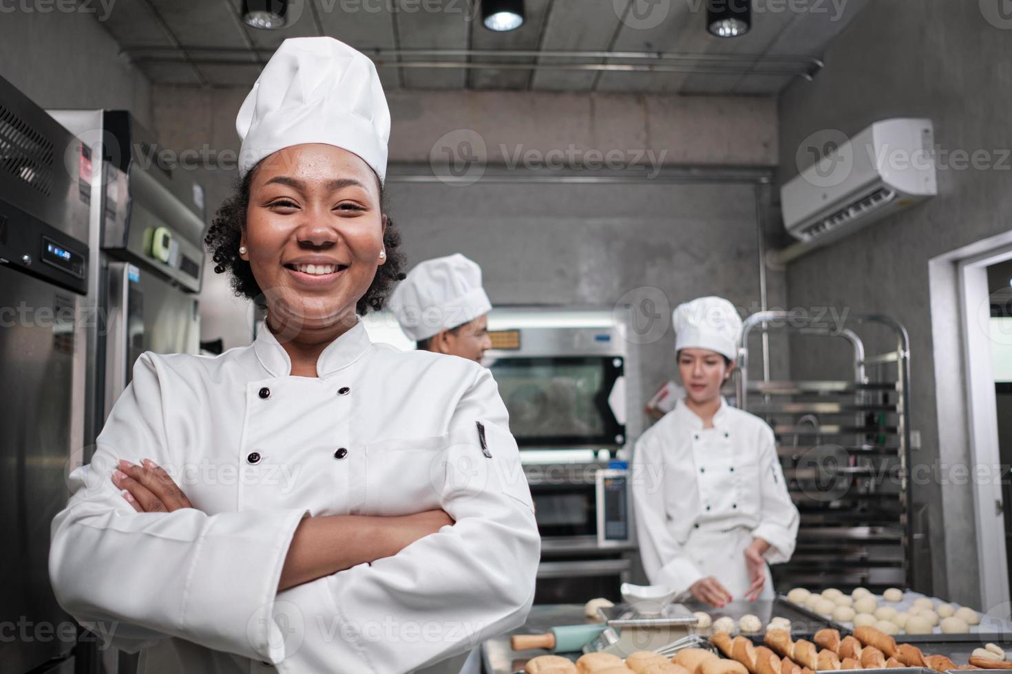 Young African American female chef in white cooking uniform looks at camera, arms crossed and cheerful smile with food professional occupation, commercial pastry culinary jobs in a restaurant kitchen. photo