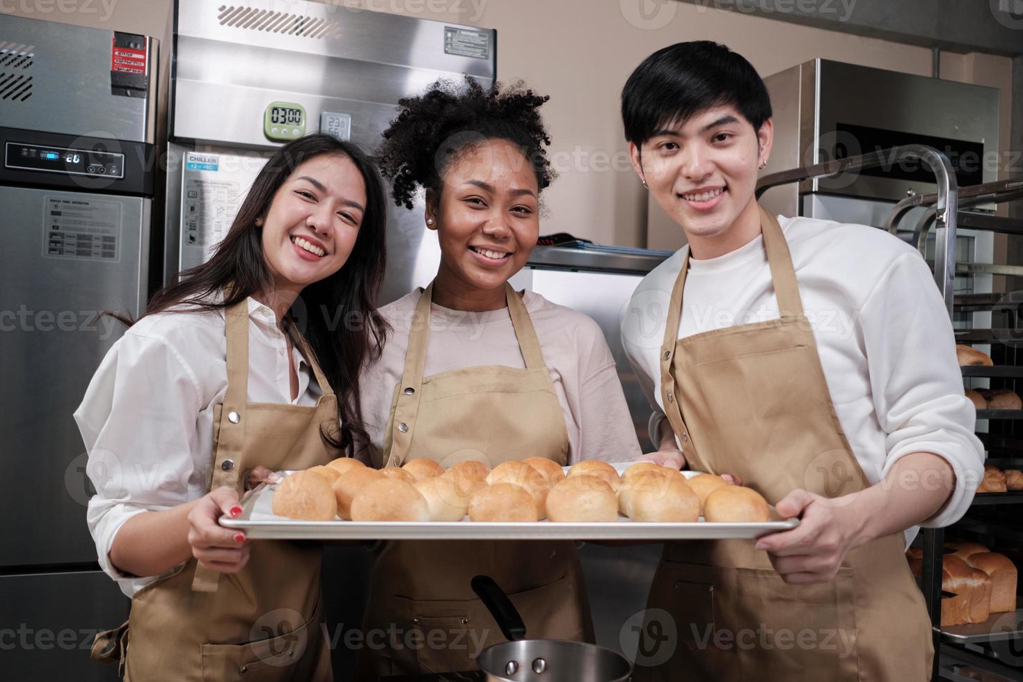 Portrait of three pastry startup partners looking at camera with a cheerful smile and proud with tray of bread in kitchen. A friend and founder of bakery foods and fresh daily bakery small business. photo