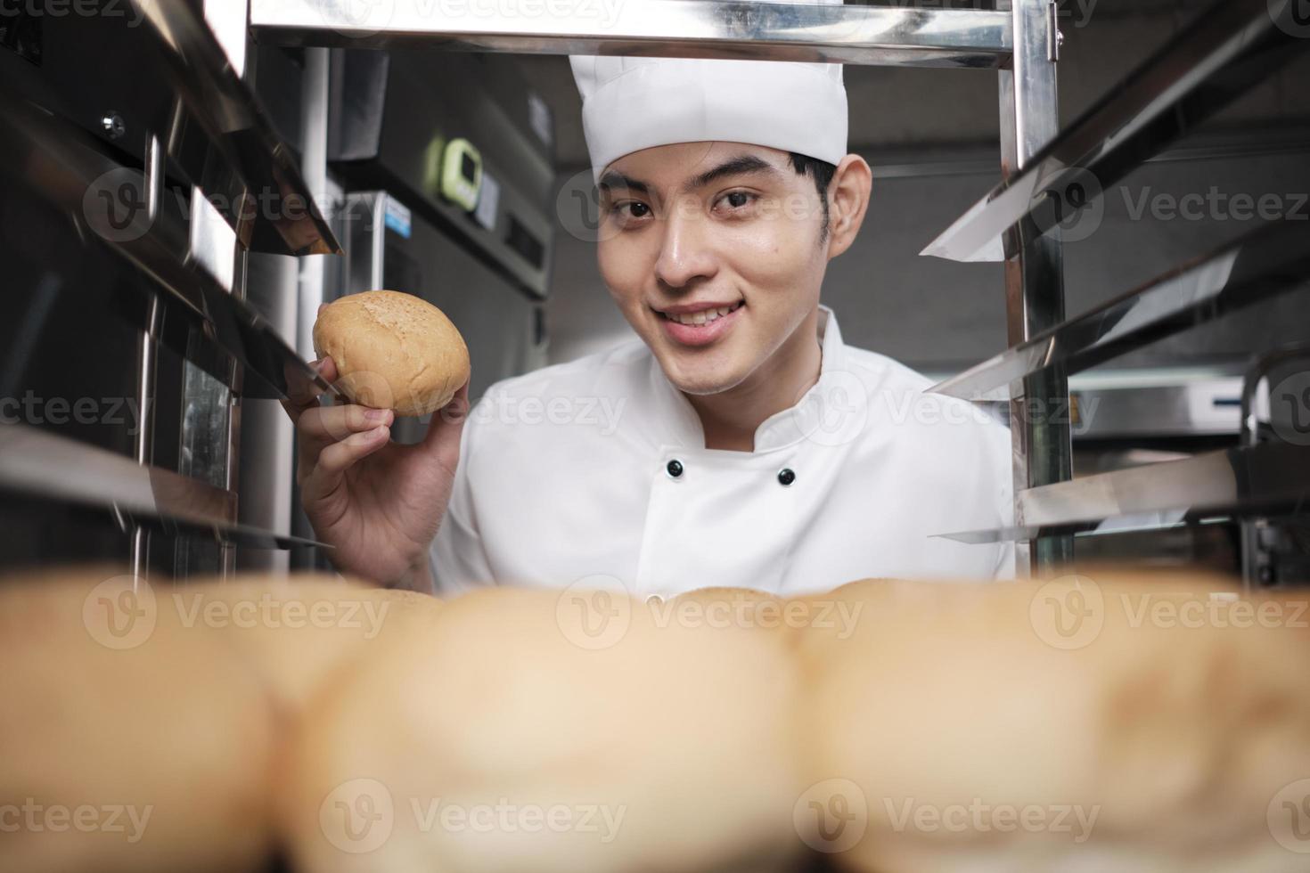 Young Asian male chef in white cook uniform and hat showing tray of fresh tasty bread with a smile, looking at camera, happy with his baked food products, professional job at stainless steel kitchen. photo