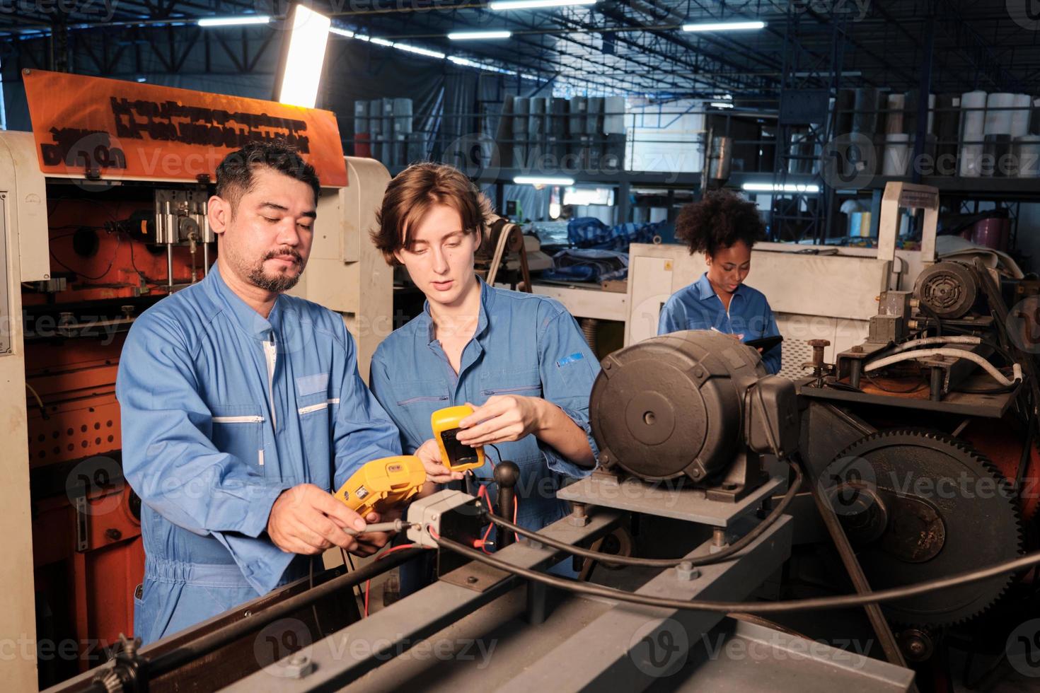 Asian male and White female engineer workers in safety uniforms work by inspecting machines' voltage current, checking, and maintaining at manufacturing factory, electric system service occupations. photo