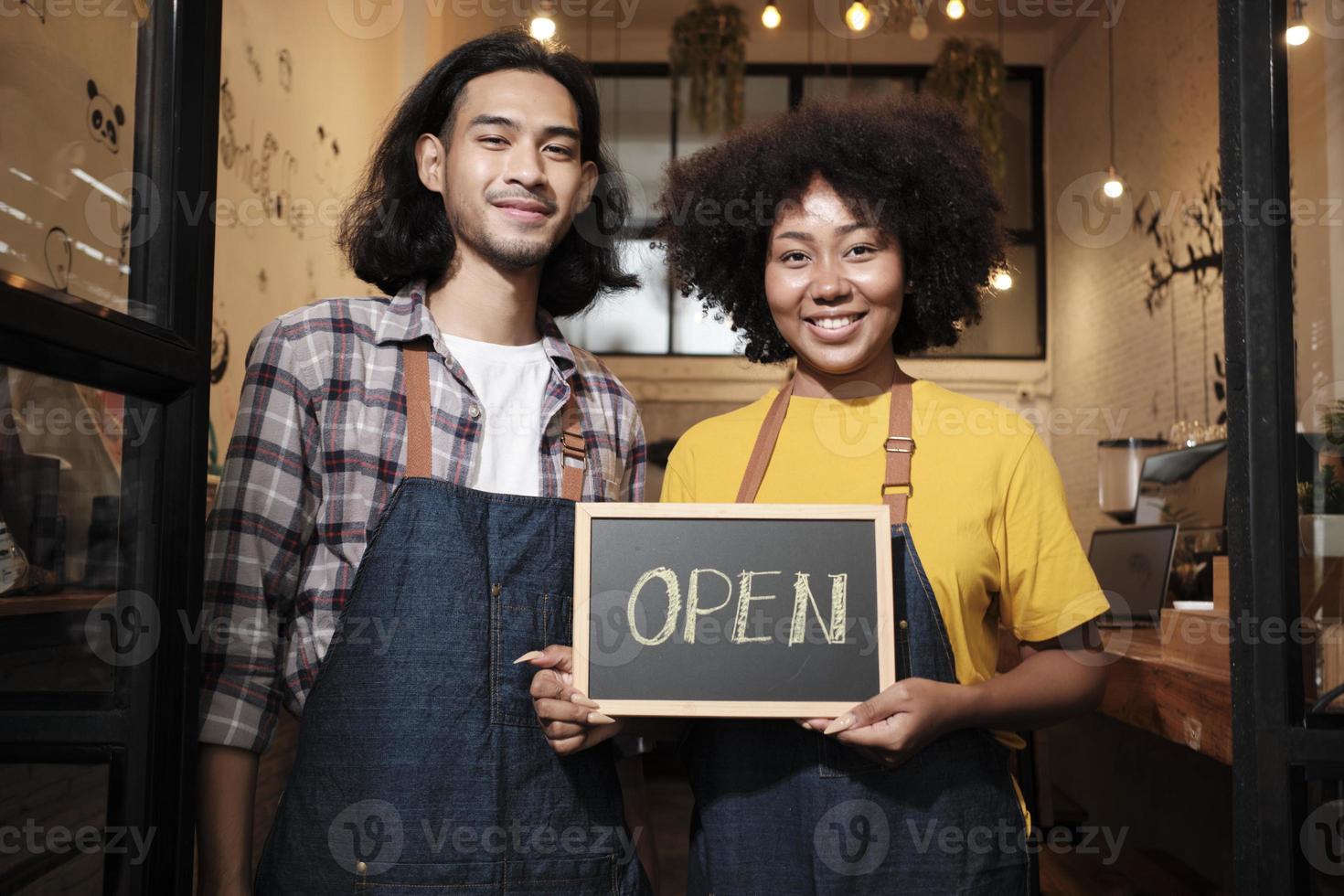 Two young startup barista partners with aprons stand at casual cafe door, letters on board and show open sign, happy and cheerful smiles with coffee shop service jobs, and new business entrepreneurs. photo