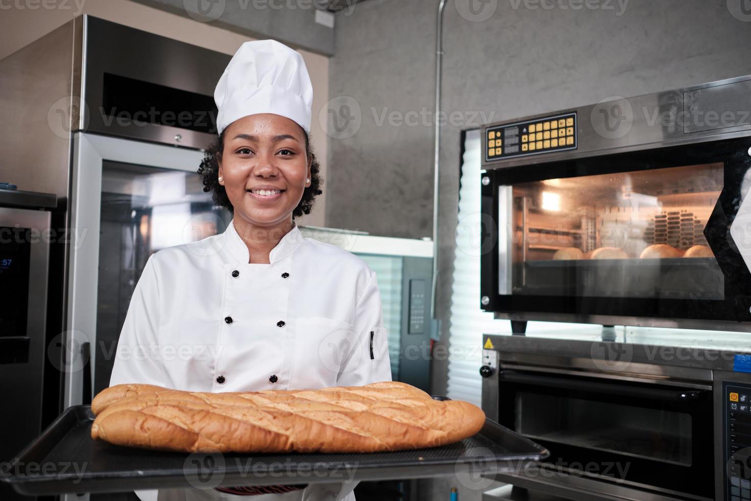 Portrait of African American female chef in white cooking uniform looking at camera with cheerful smile and proud with tray of baguette in kitchen, pastry foods professional, fresh bakery occupation. photo