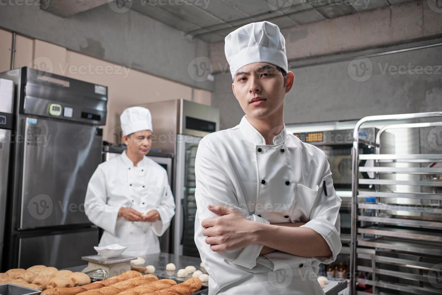 Portrait of young Asian male chef in white cooking uniform looking at the camera, arms crossed with confidence, foods professional occupation, commercial pastry culinary jobs in a restaurant kitchen. photo