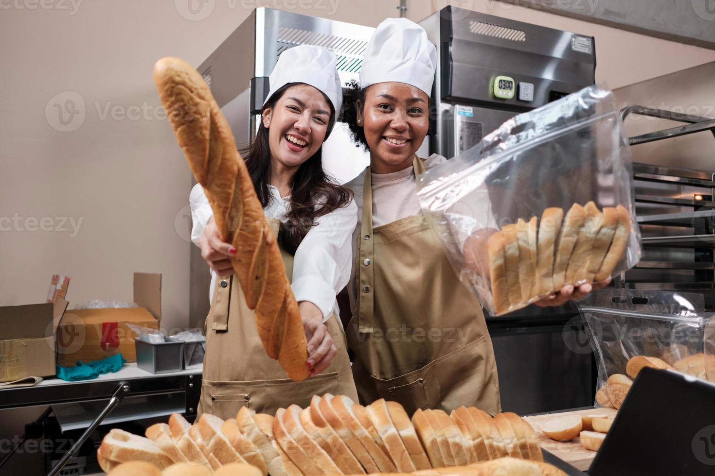 Portrait of young female chefs in apron uniforms with hats looking at camera with cheerful smile and proud with bread in kitchen. Friend and partner of bakery foods and fresh daily bakery occupation. photo