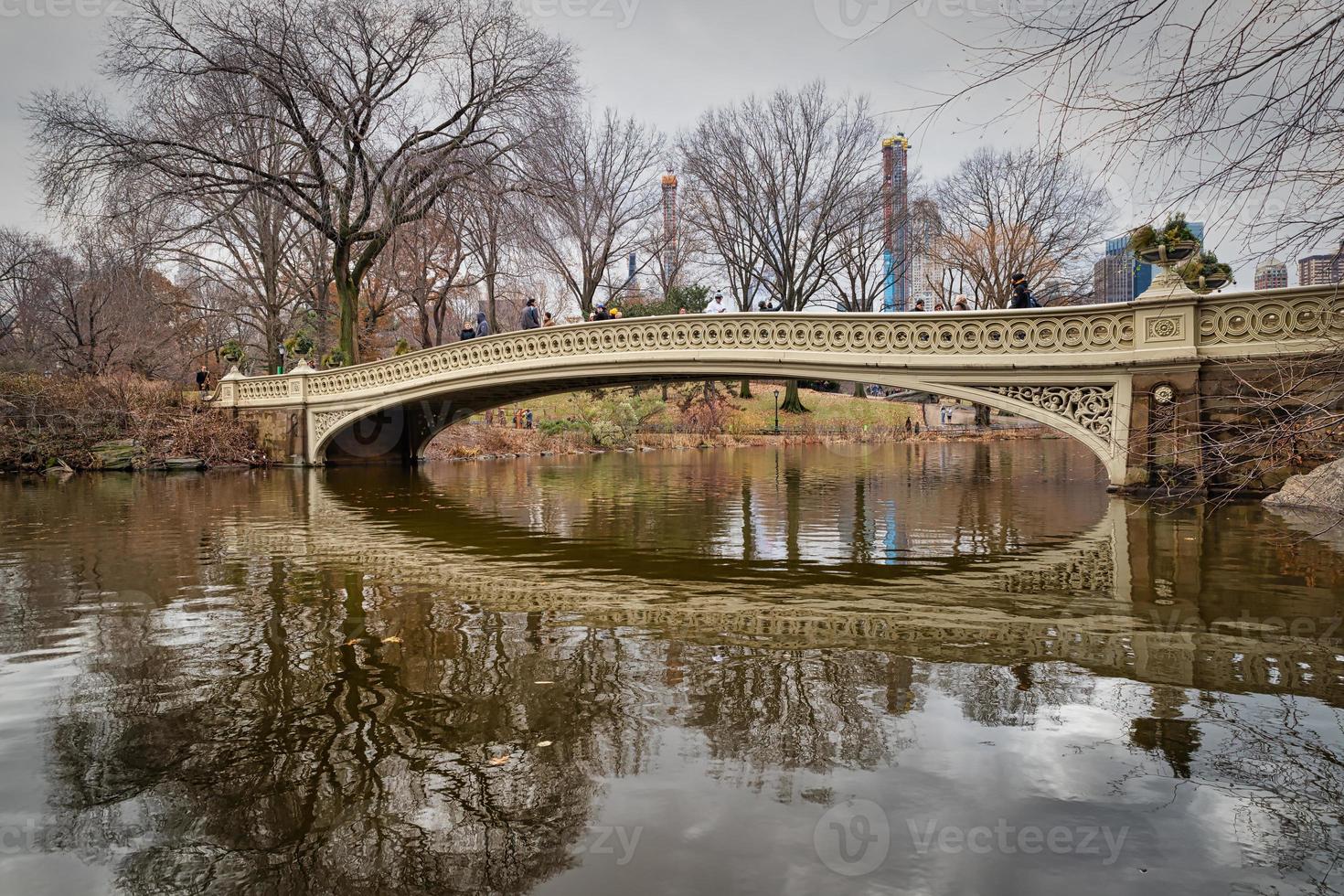 The bow bridge in central park, New York city daylight view with reflection in water , clouds, trees and Manhattan skyline in background photo