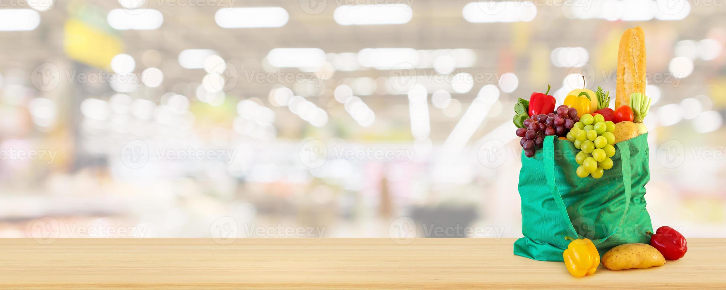 Fresh fruits and vegetables in reusable green shopping bag on wood table with supermarket grocery store blurred defocused background photo