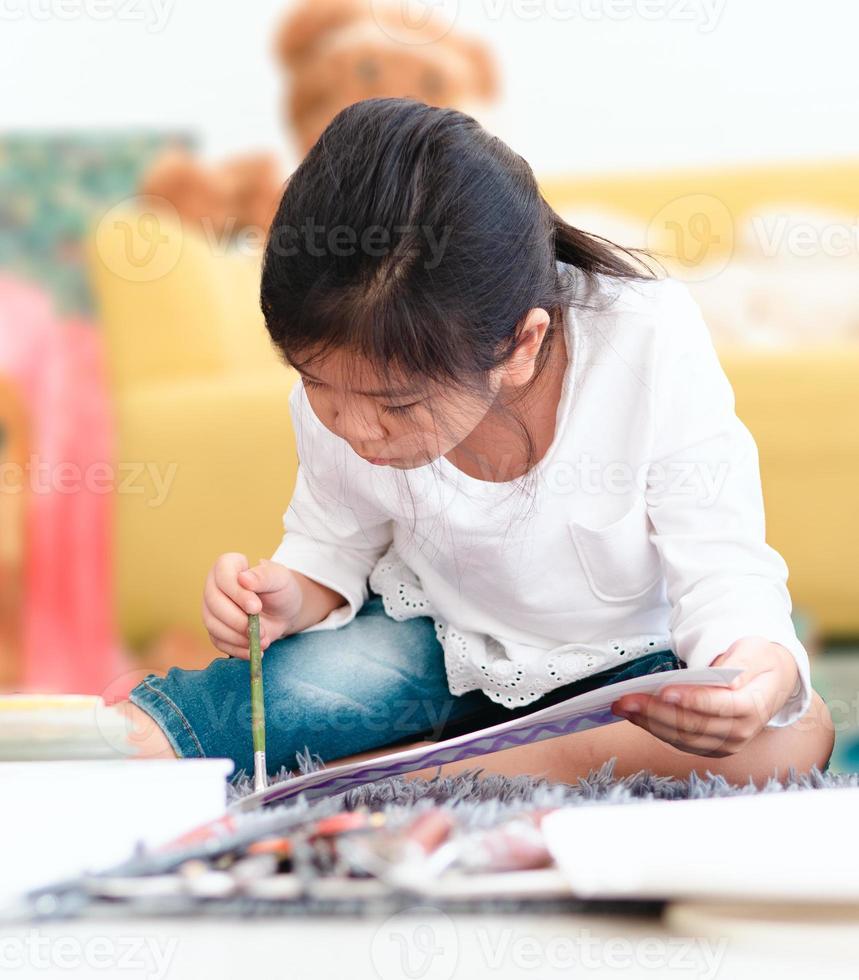 niño pequeño sentado en el suelo dibujando bocetos en papel. adorable joven artista disfruta pintando la tarea. niño asiático juguetón aprende a construir ideas creativas, inspiración, imaginación a través del dibujo foto
