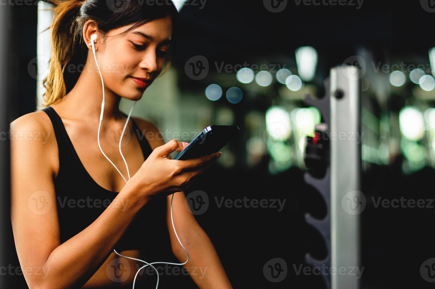 chica en el gimnasio haciendo ejercicio usa un teléfono celular escuchando música con auriculares blancos y usando un temporizador digital de latidos del corazón. ejercicio sistemático sus vacaciones relajación foto