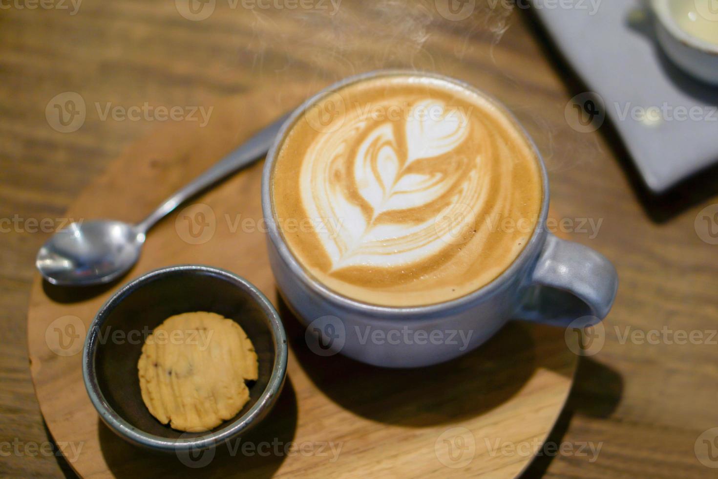 Top view of Late coffee and late art in blue ceramic coffee cup with a cookie in a black ceramic bowl on wooden plate and table in the coffee shop. photo