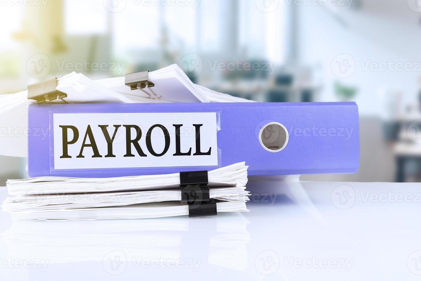 Payroll, the term is written on a folder of documents in trendy purple, lying on a stack of documents on an office table against the backdrop of an office with a soft blurred background. photo