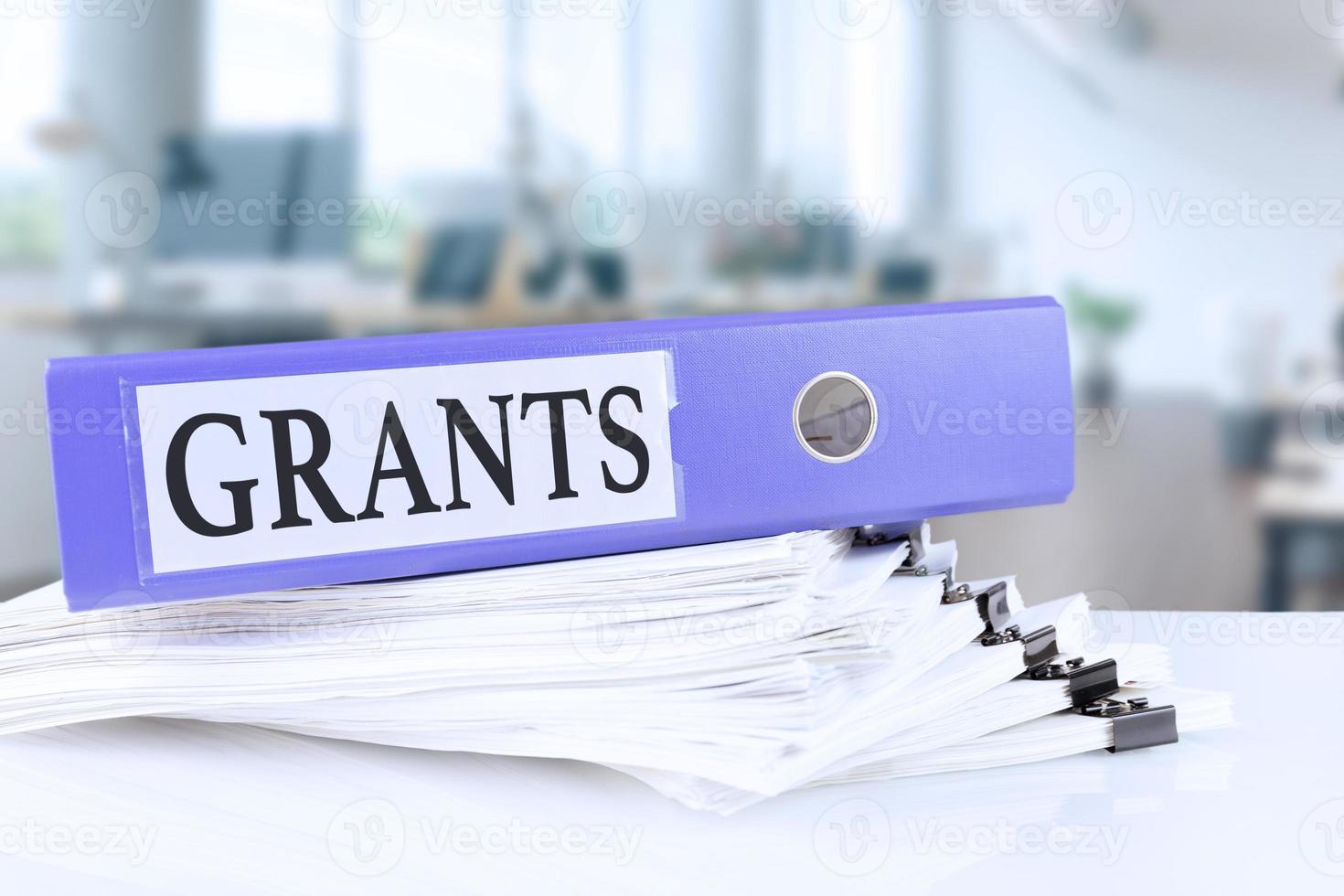 Grants, the term is written on a folder of documents in trendy purple, lying on a stack of documents on an office table against the backdrop of an office with a soft blurred background. photo