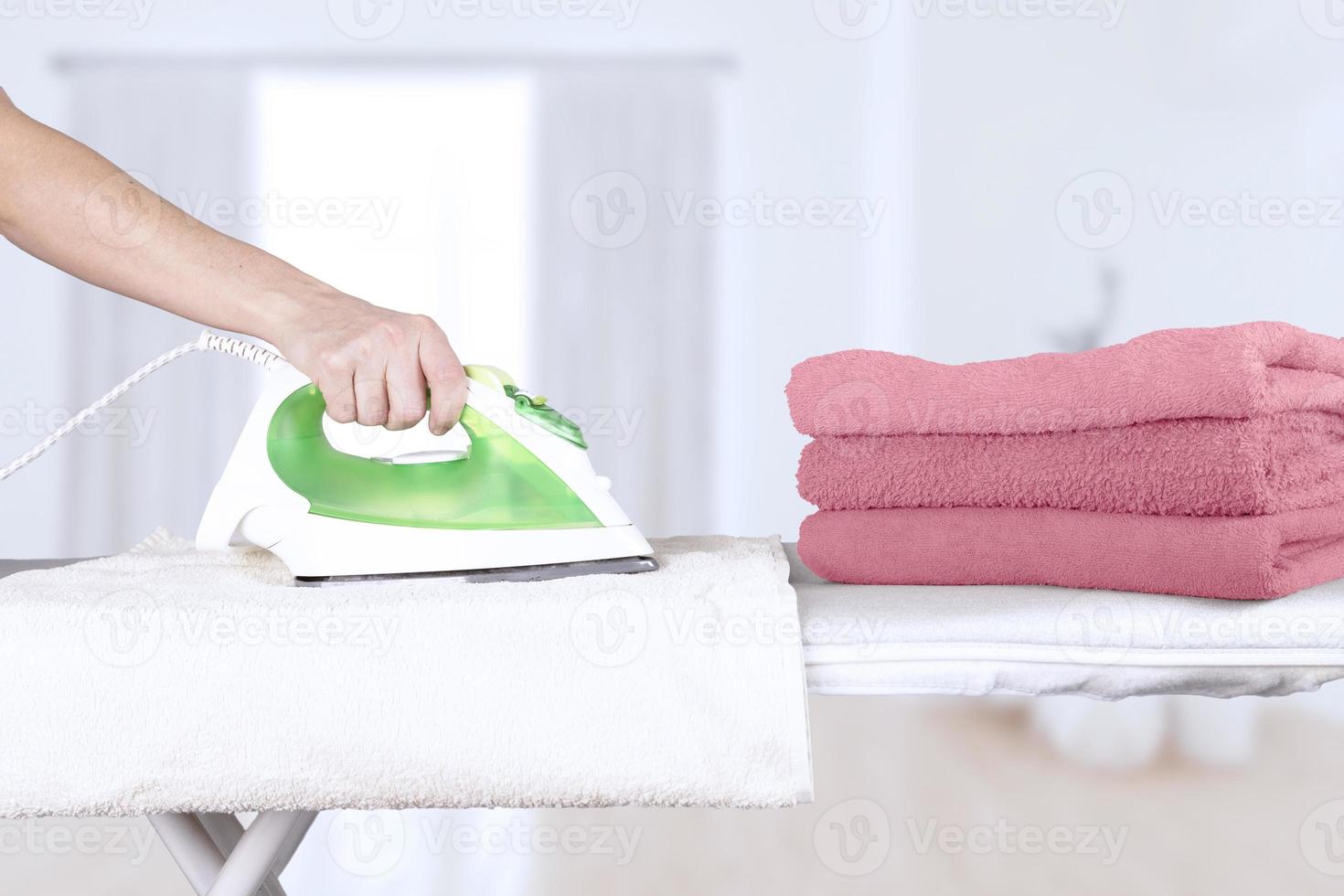 mujer caucásica, esposa, madre, ama de casa, ama de llaves, criada planchando toallas de baño en tonos rosas, plancha eléctrica en tabla de planchar. fondo interior de la casa. enfoque suave. foto