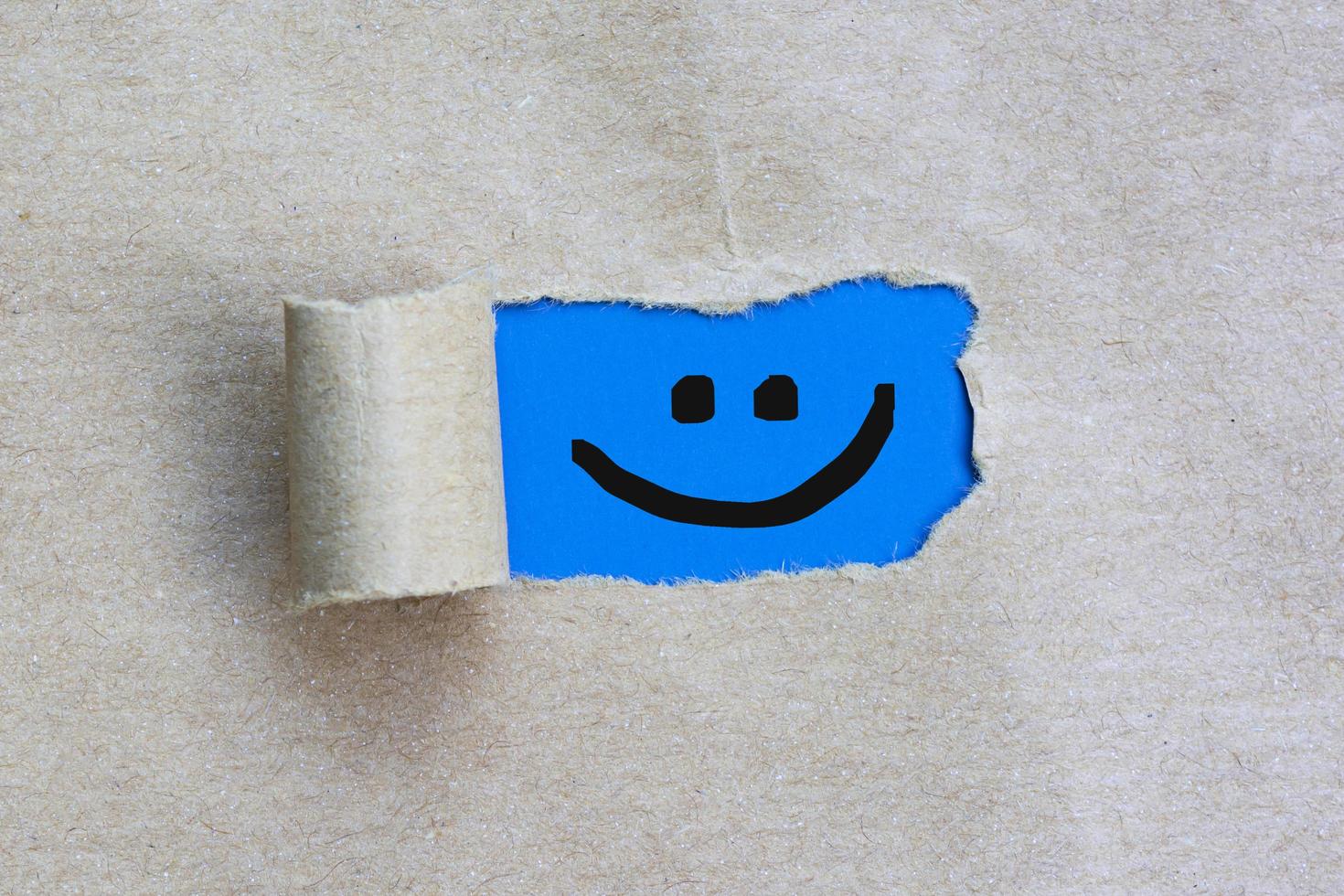 Smiley face on torn hole in the sheet of brown paper on blue background. photo