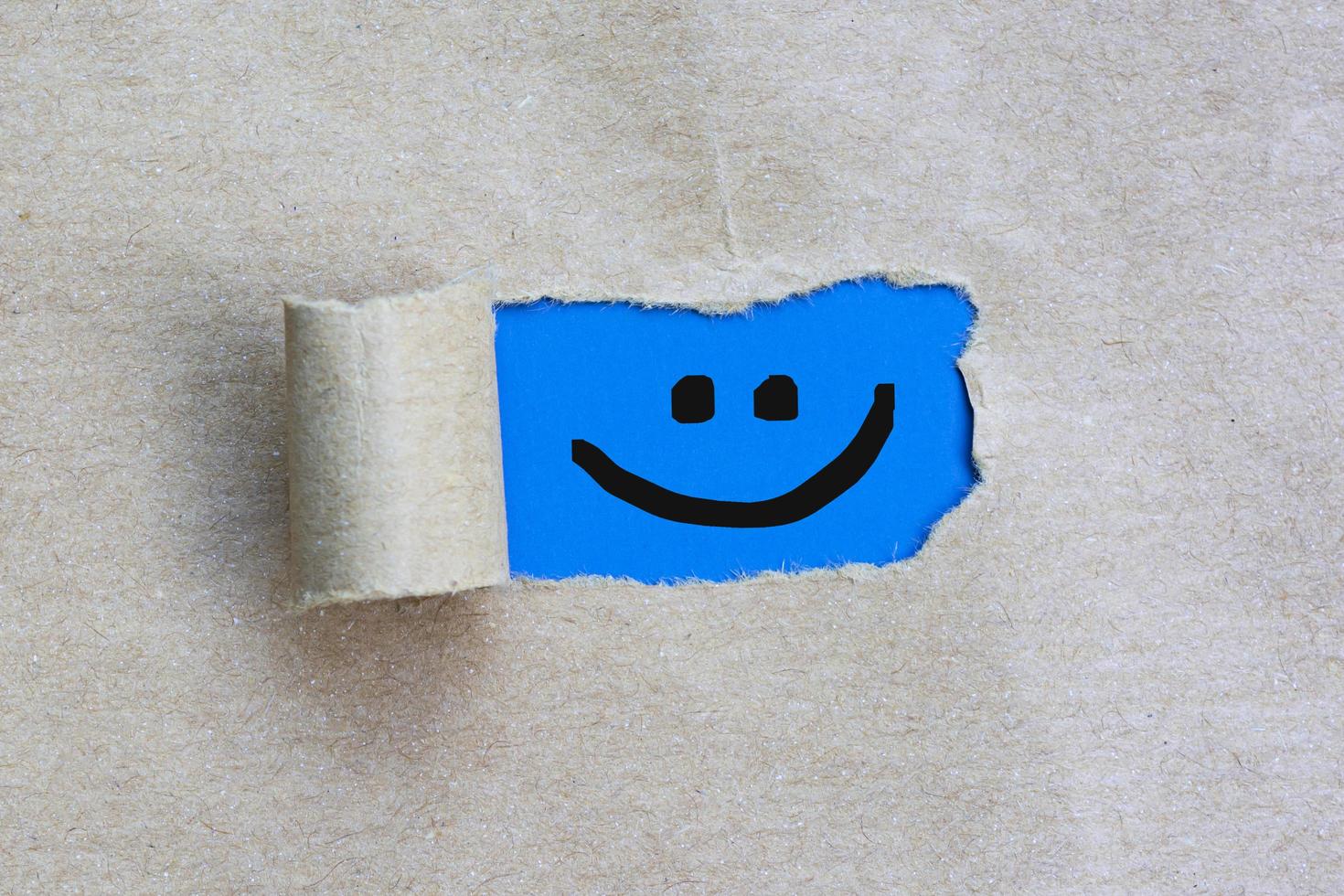 cara sonriente en un agujero rasgado en la hoja de papel marrón sobre fondo azul. foto