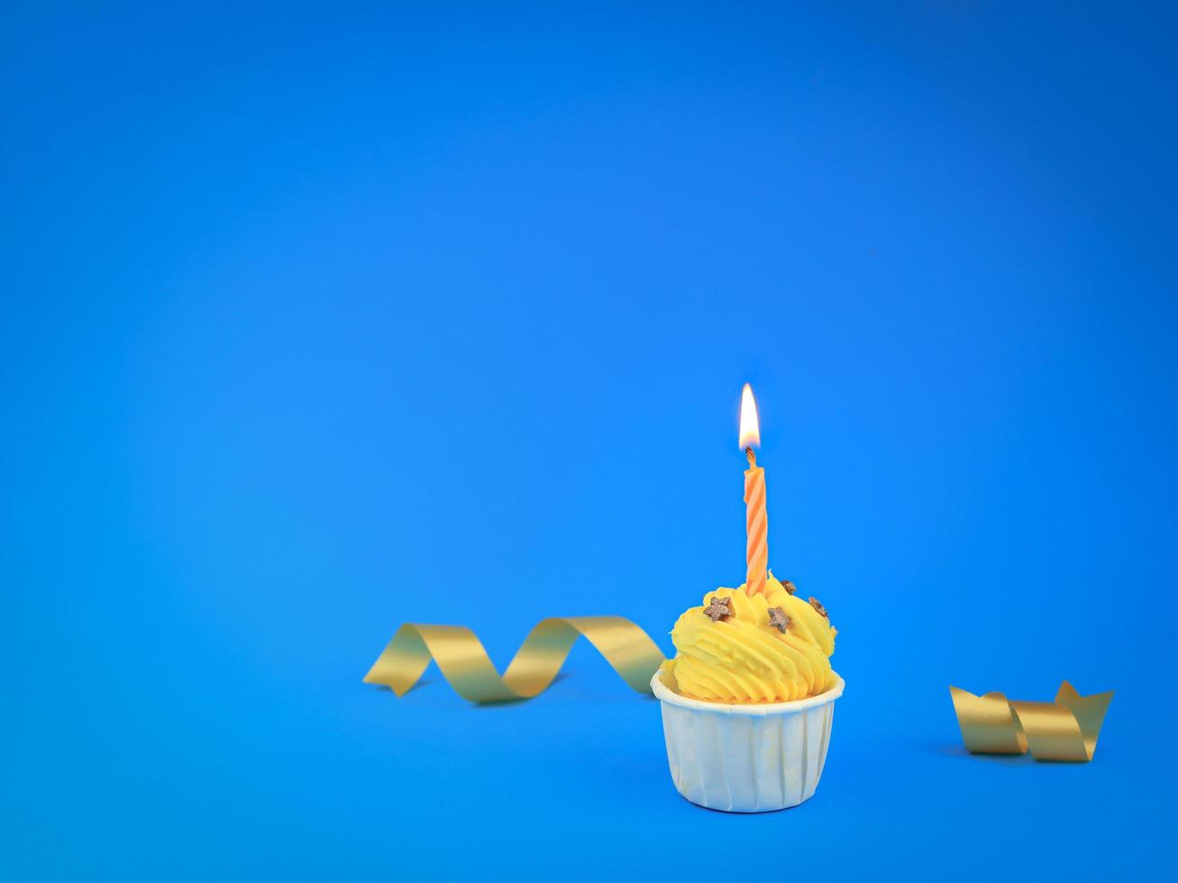 Sweet bright yellow cupcake with bow candle on blue background with copy space. Happy birthday party photo