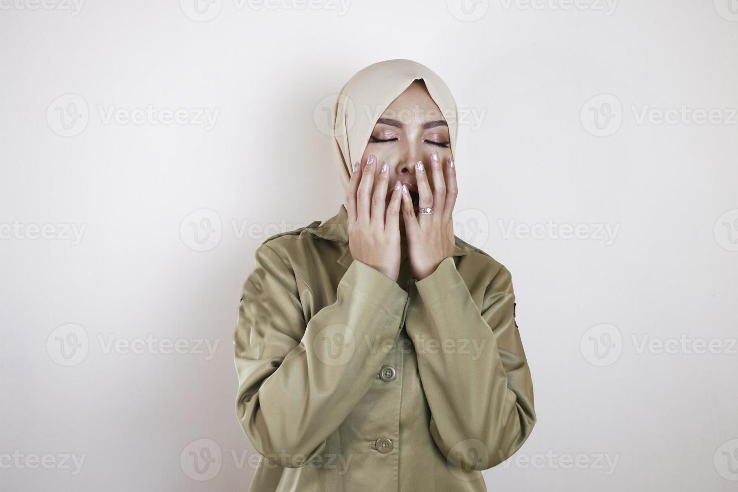 Portrait of sleepy Asian Muslim woman wearing a brown uniform and hijab, covering her mouth with a hand while yawning. photo