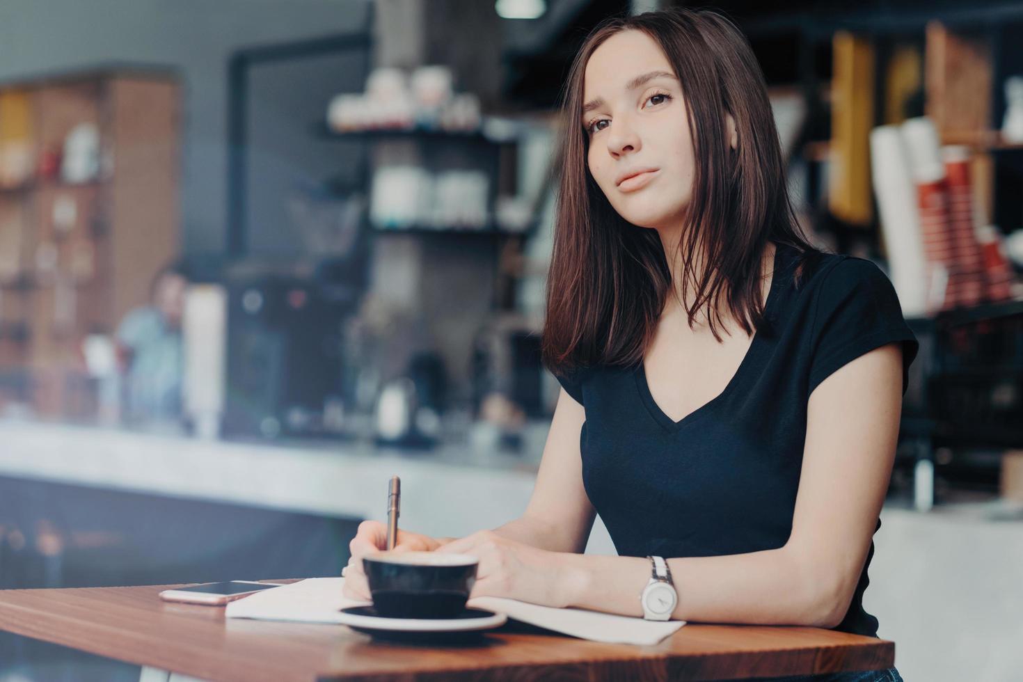 Concentrated female journalist thinks about new article, writes down information into notepad, drinks hot aromatic beverage in coffee shop, modern smart phone on table near. Working process. photo
