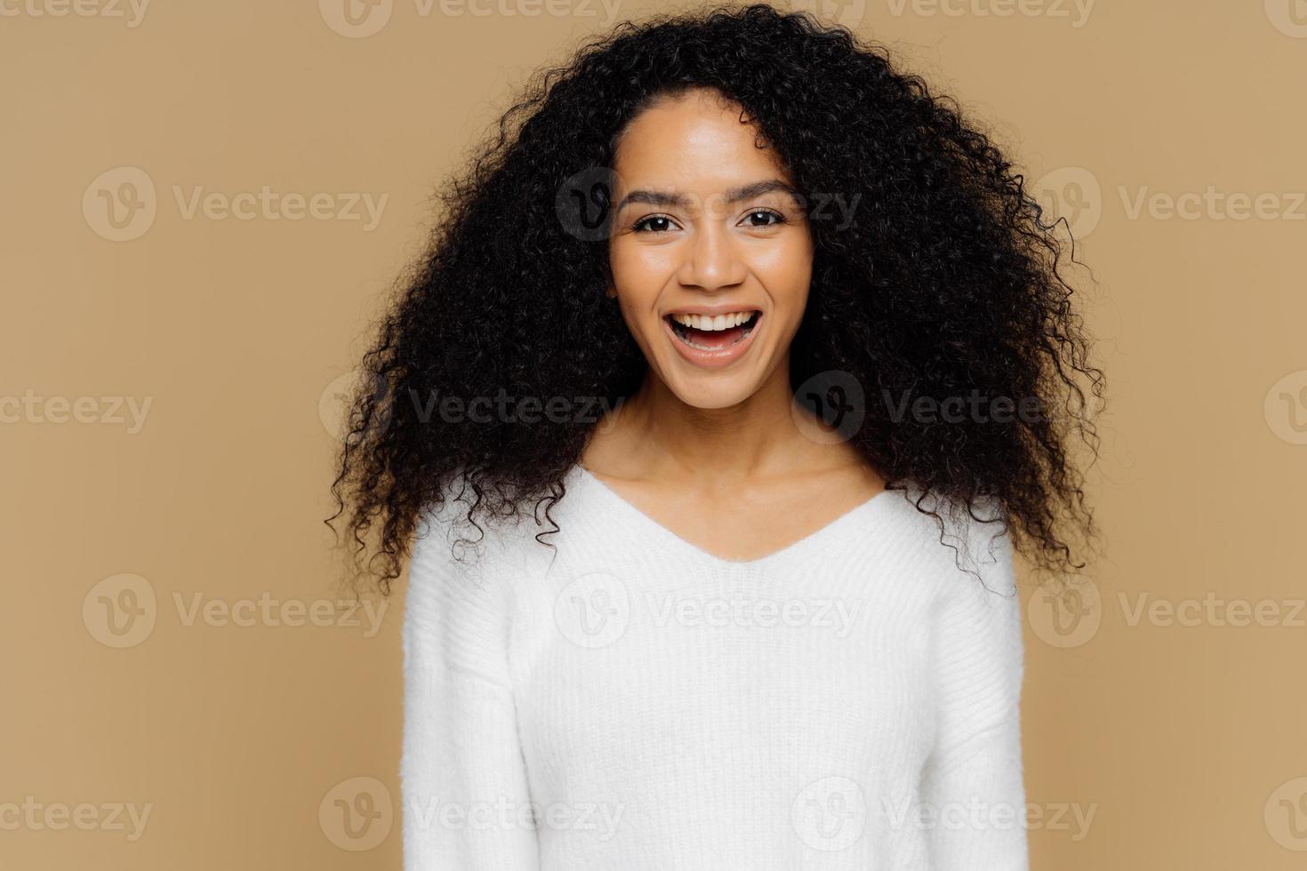 Headshot of healthy positive female model laughs happily, has bushy Afro hair, dressed in white soft jumper, glad to receive compliment from boyfriend, isolated over brown background. Emotions concept photo