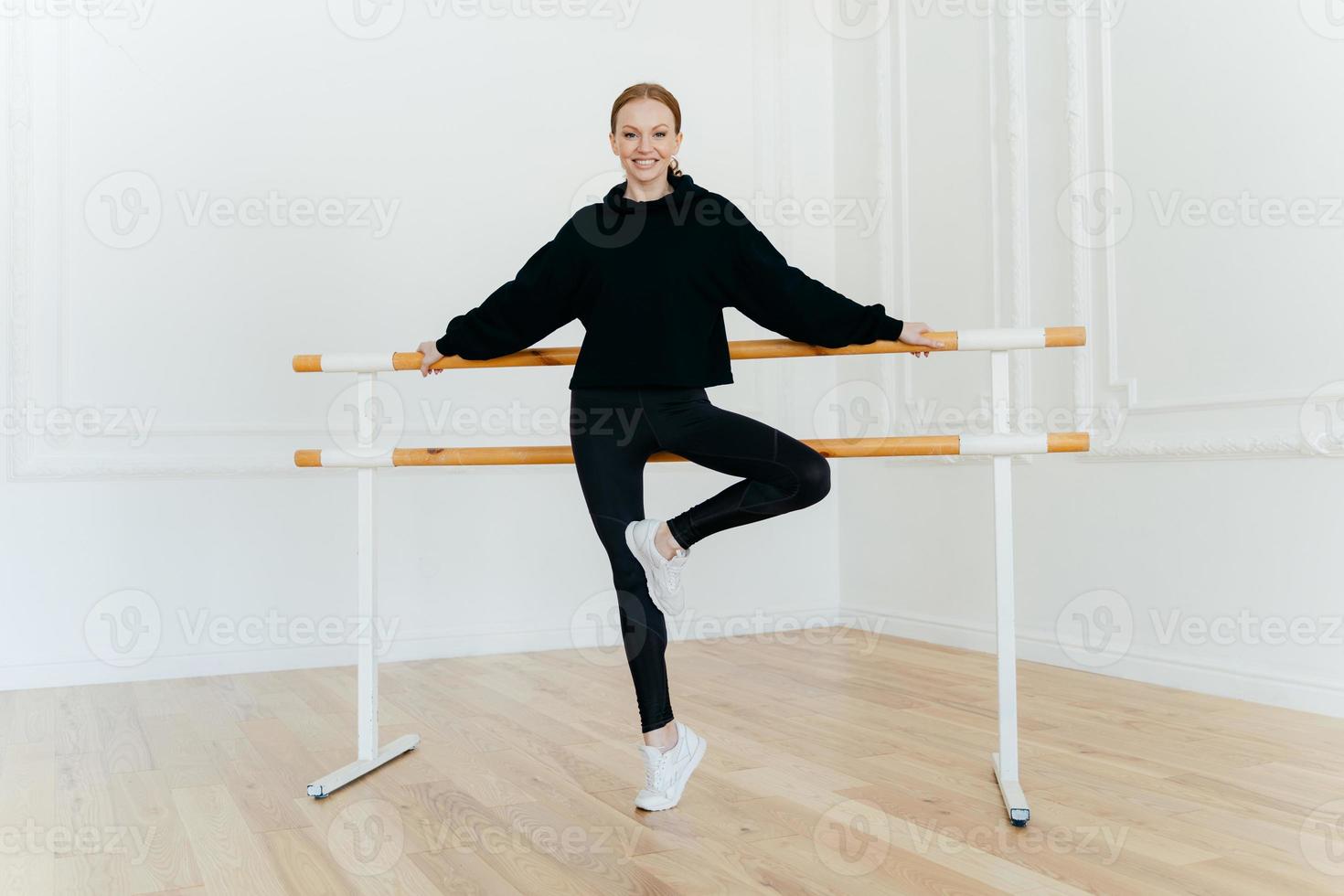 Horizontal shot of flexible young female leans on ballet barre, wears black costume and white sportshoes, smiles positively, poses in dancing hall. Ballet instructor demonstrates different exercises photo