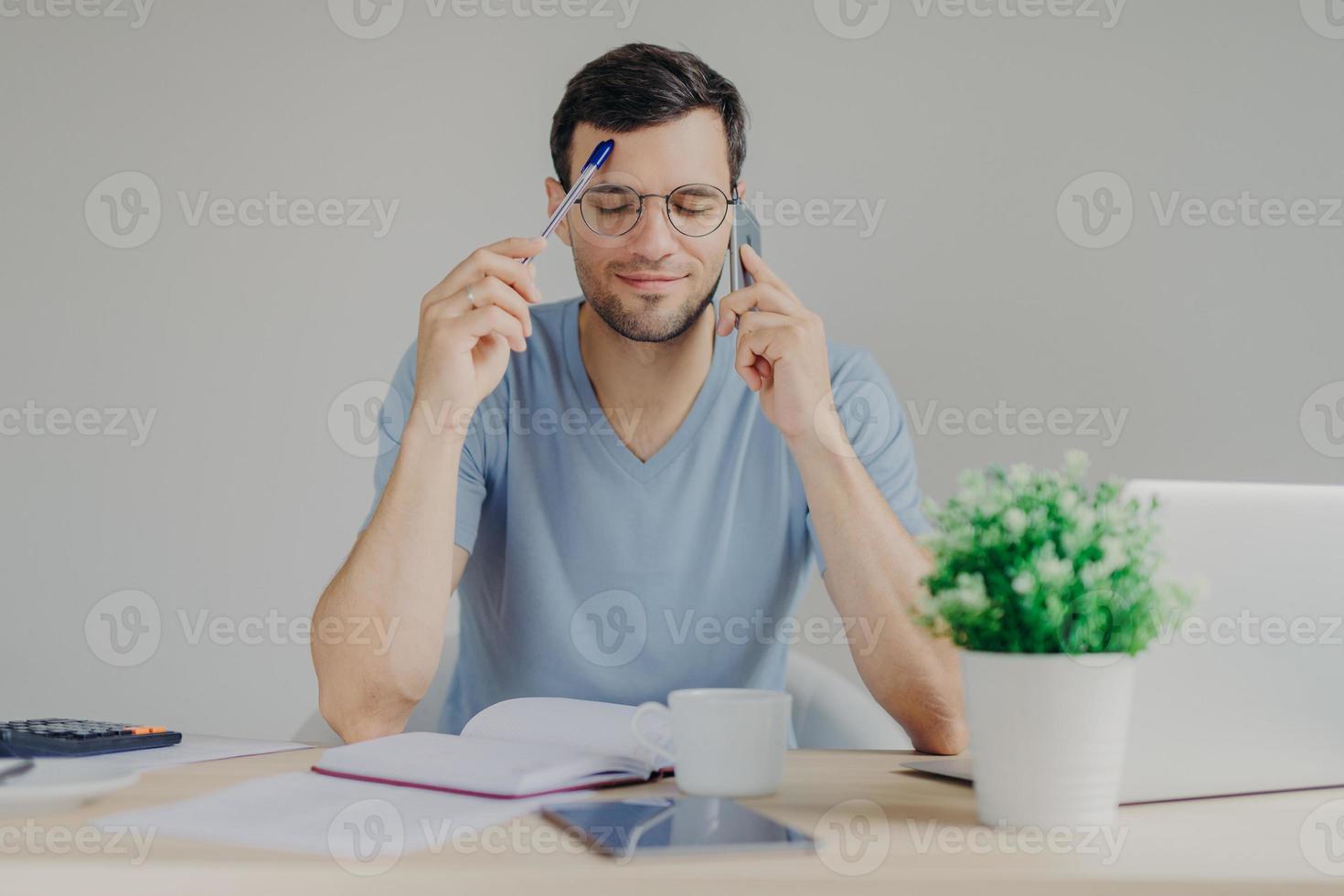 Busy male works on organizational issues, speaks with business partner via cell phone, keeps eyes closed as tries to remember necessary information and write in diary. People and work concept photo